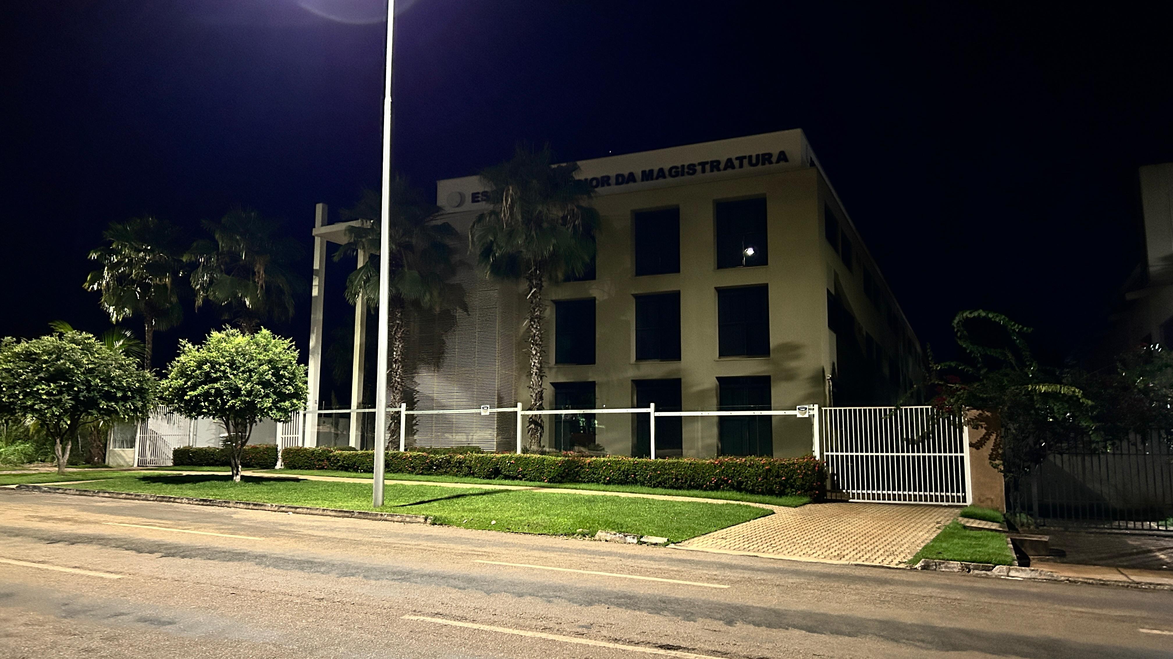 Fachada do prédio da Esmat às escuras, com vegetação ao redor e uma rua na frente.