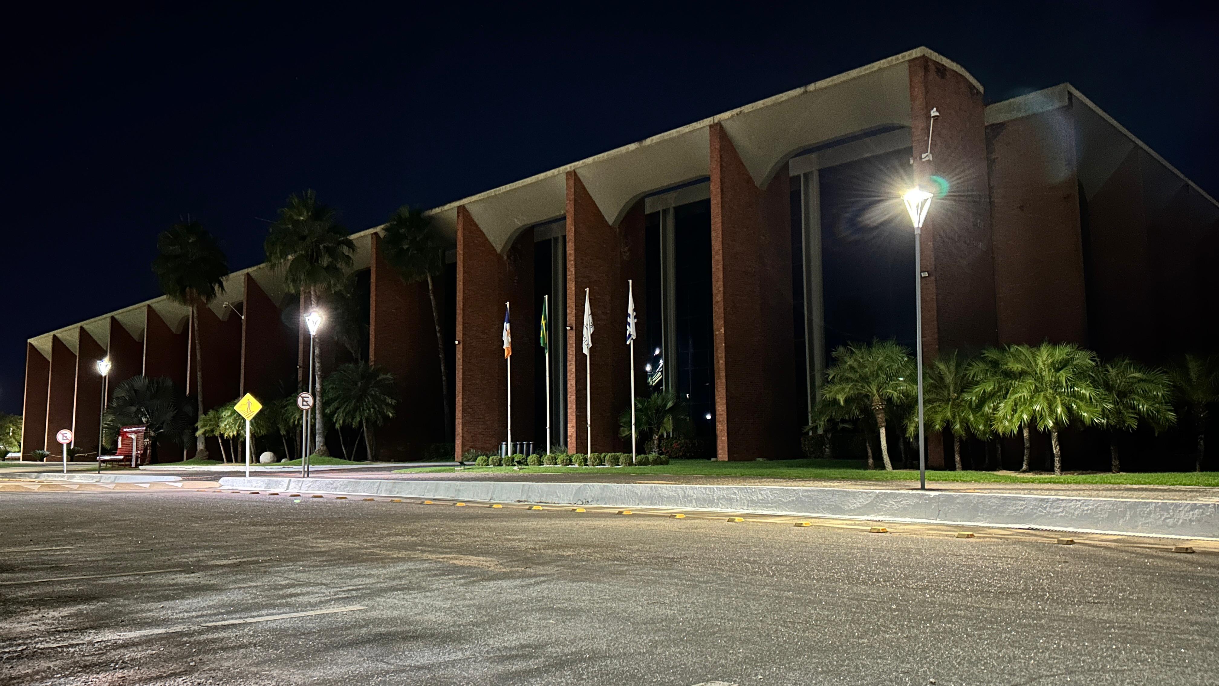 Fachada do prédio do Tribunal de Justiça com as luzes apagadas, à frente aparecem uma calçada, jardim e uns postes com as luminárias ligadas. 