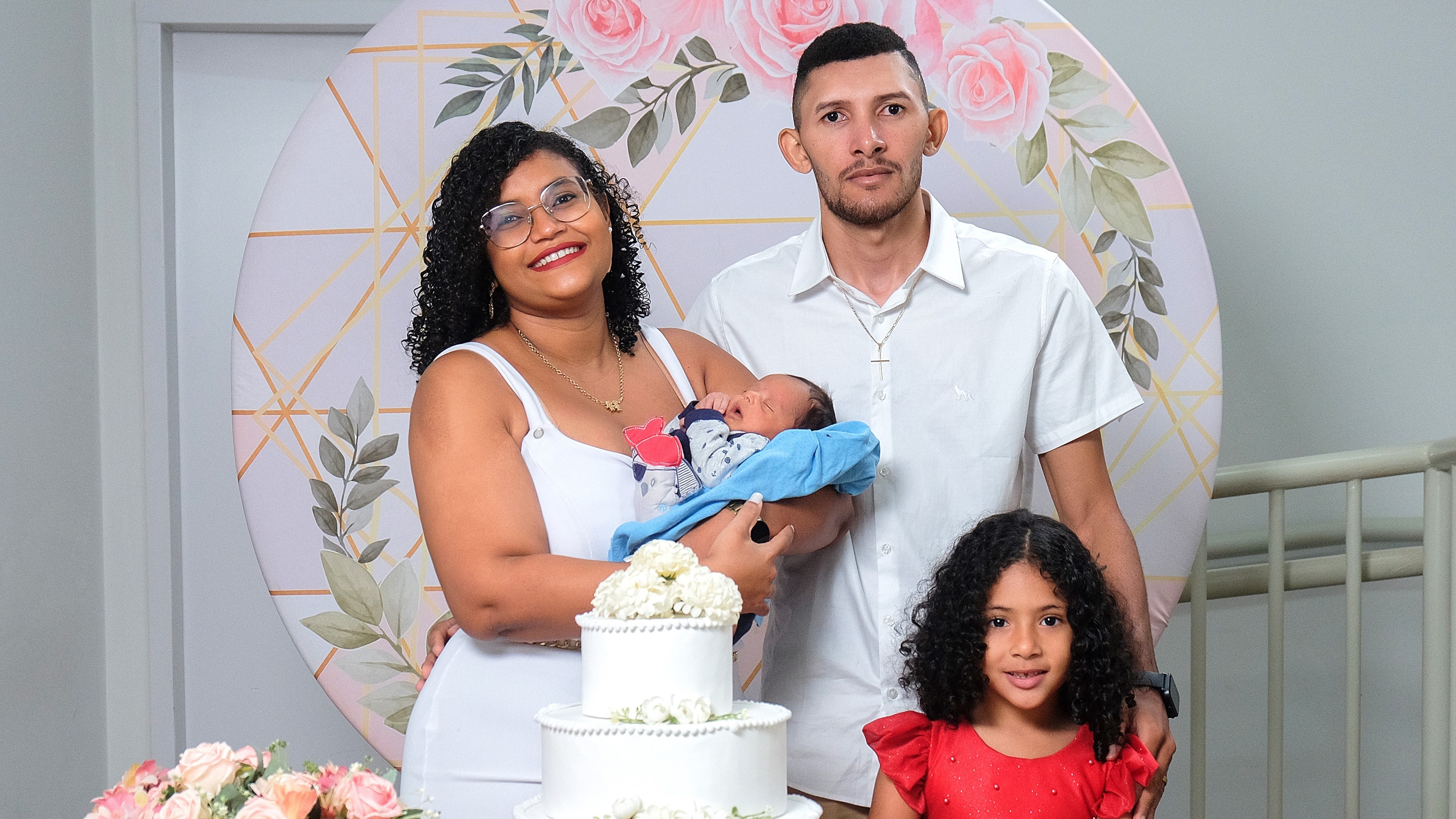 No centro da imagem, o casal de noivos posa sorridente, vestidos com trajes tradicionais: a noiva em um vestido branco e o noivo de terno. A noiva segura carinhosamente um bebê no colo, enquanto à frente do casal tem uma menina de cabelos longos cacheados e vestido vermelho. Em destaque, bem ao centro da imagem, está um bolo de casamento decorado, simbolizando a celebração do amor e da família.