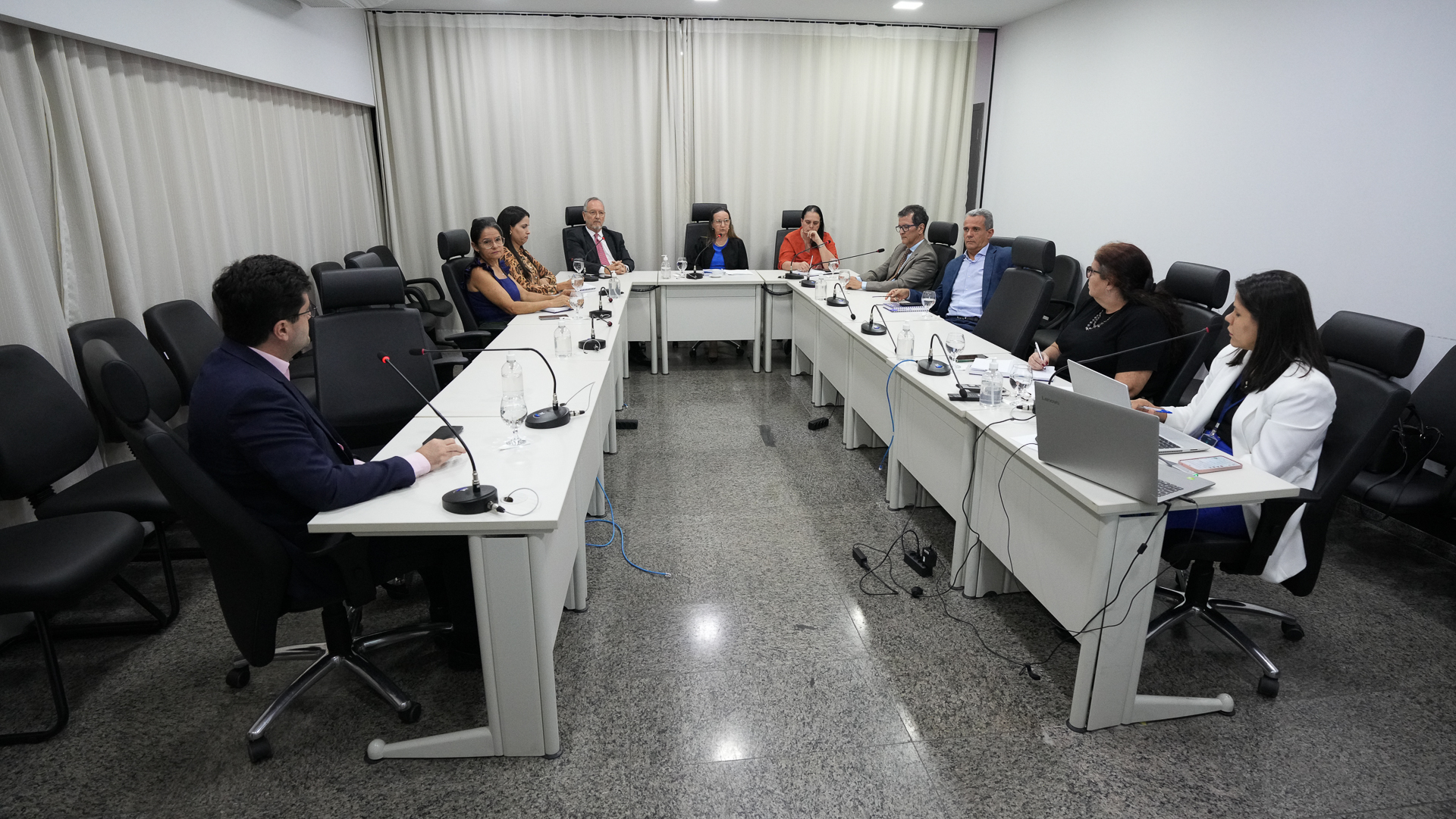 A imagem mostra uma reunião formal do Tribunal de Justiça do Tocantins (TJTO). O ambiente é bem iluminado, com luzes de teto brancas, cortinas claras e paredes neutras. Dez pessoas estão sentadas ao redor de uma mesa em formato de “U”, com microfones, blocos de anotações e copos de água à disposição, o que indica um encontro institucional ou estratégico. Entre os participantes, há magistradas e magistrados, além de servidores, todos atentos à discussão.