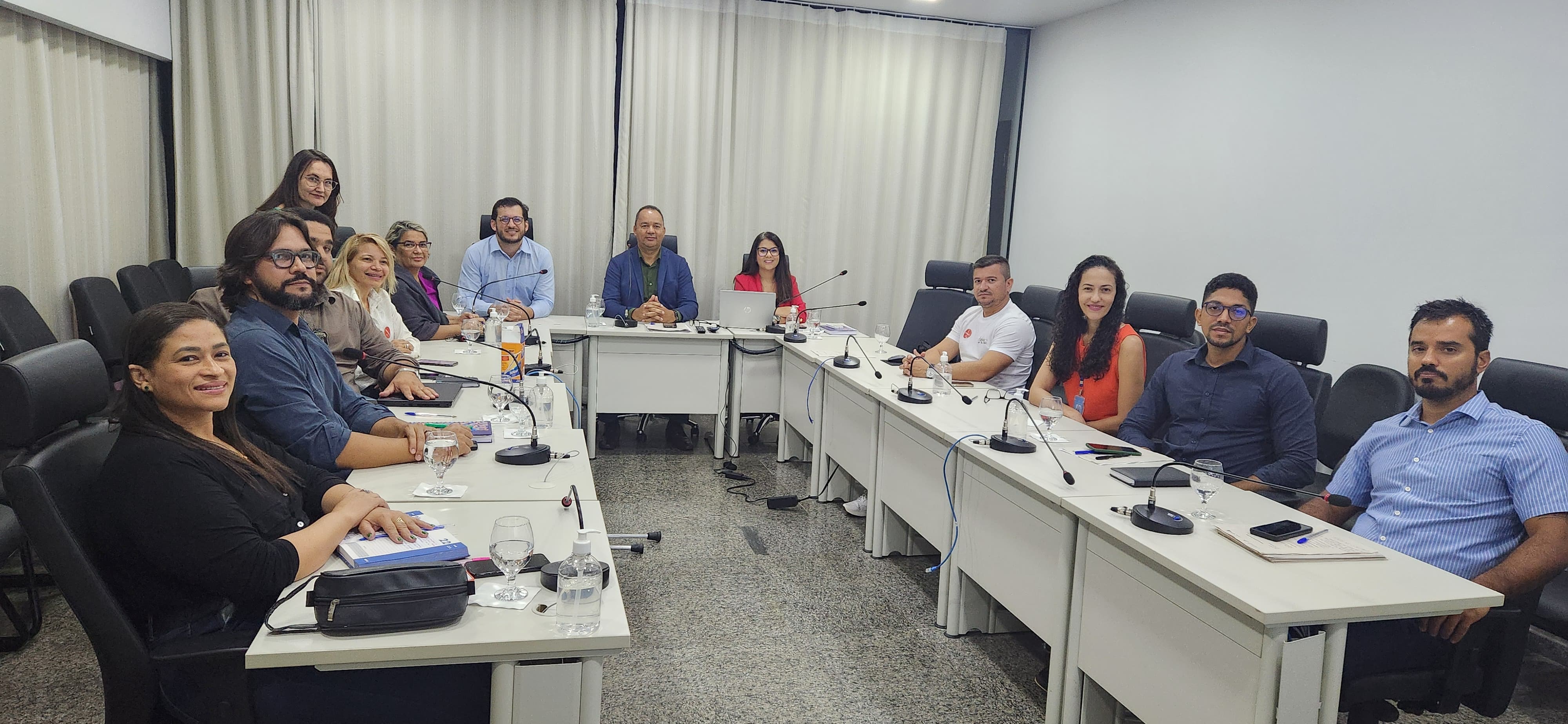 Imagem mostra uma sala de reuniões e várias pessoas sentadas; na imagem aparecem mesas brancas em formato em "U"