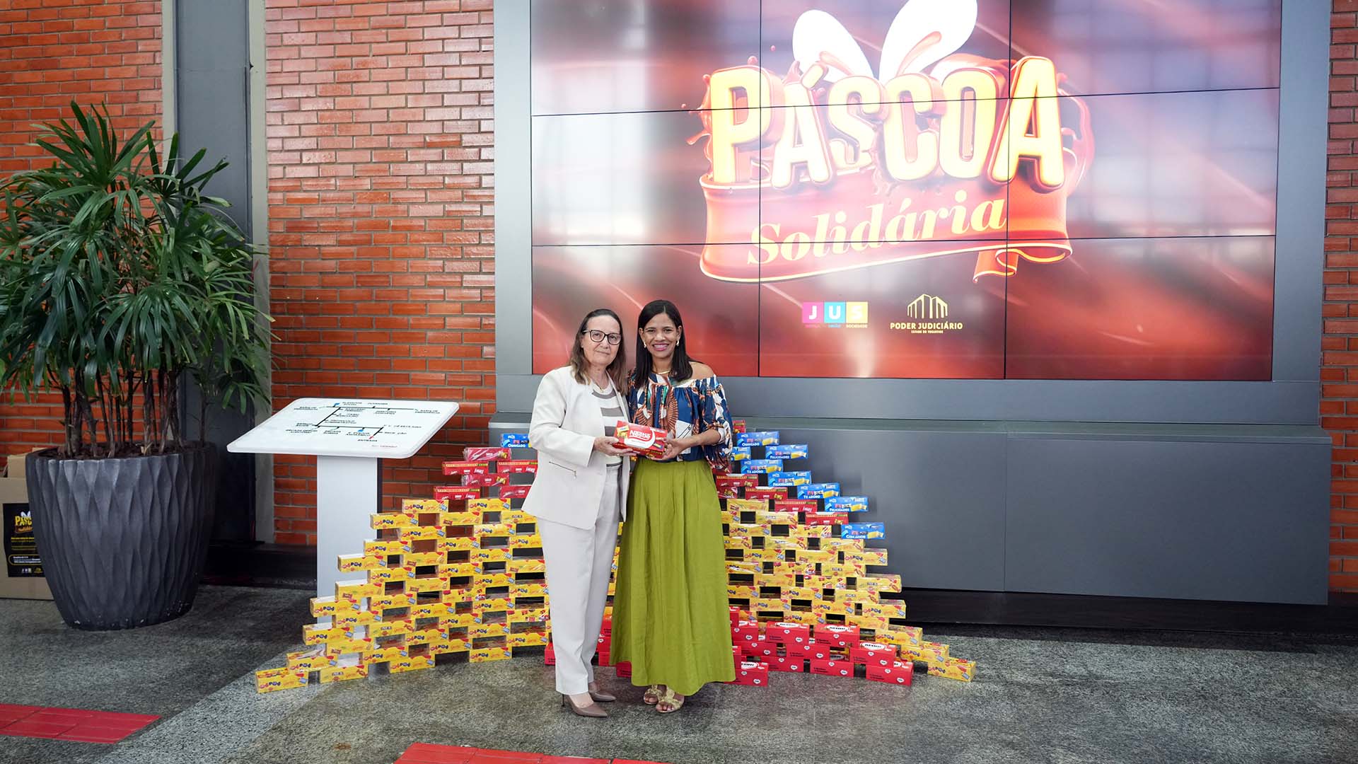 Desembargadora Maysa à esquerda e a diretora da escola estão posicionadas em frente a uma grande tela, que exibe uma mensagem de Páscoa Solidária. Elas seguram uma caixa de chocolate e ao fundo delas, no chão, aparece uma grande quantidade de caixas de chocolate empilhada.