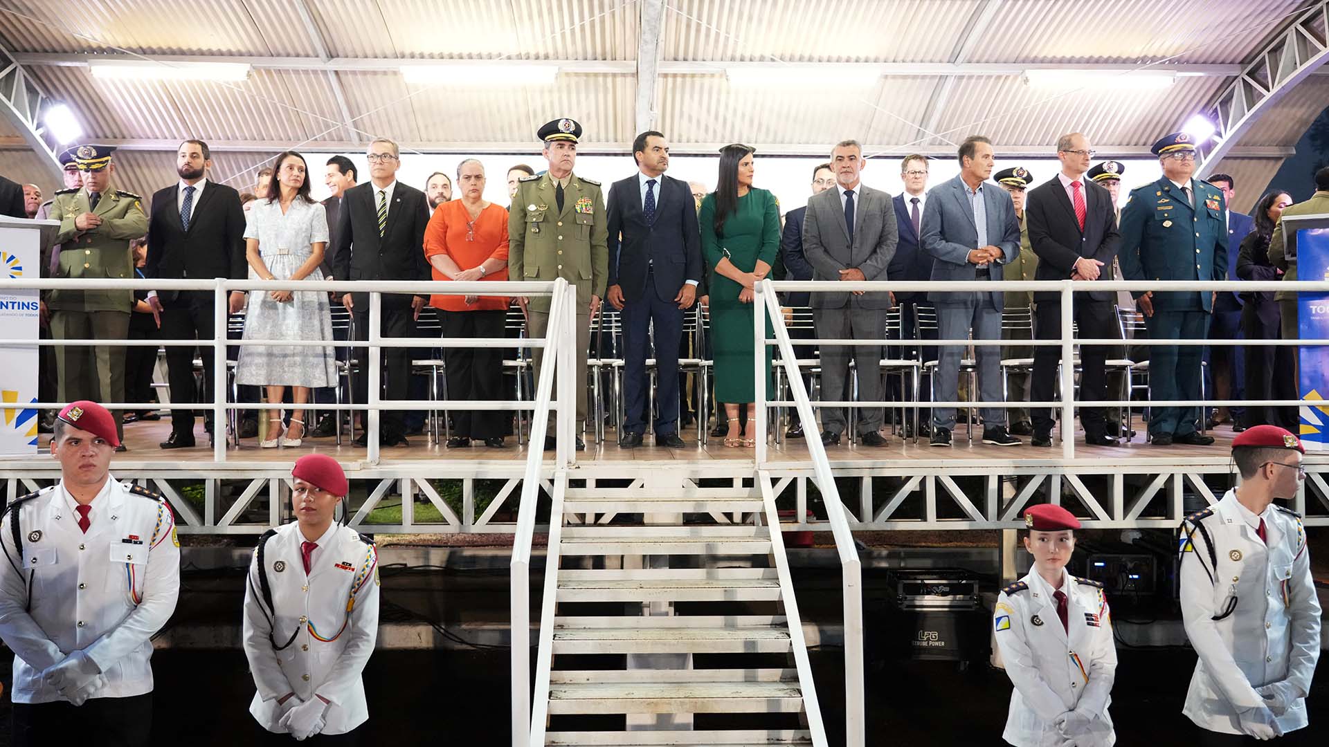  imagem mostra várias autoridades, em trajes formais, em pé, de frente, no palanque, durante cerimônia.