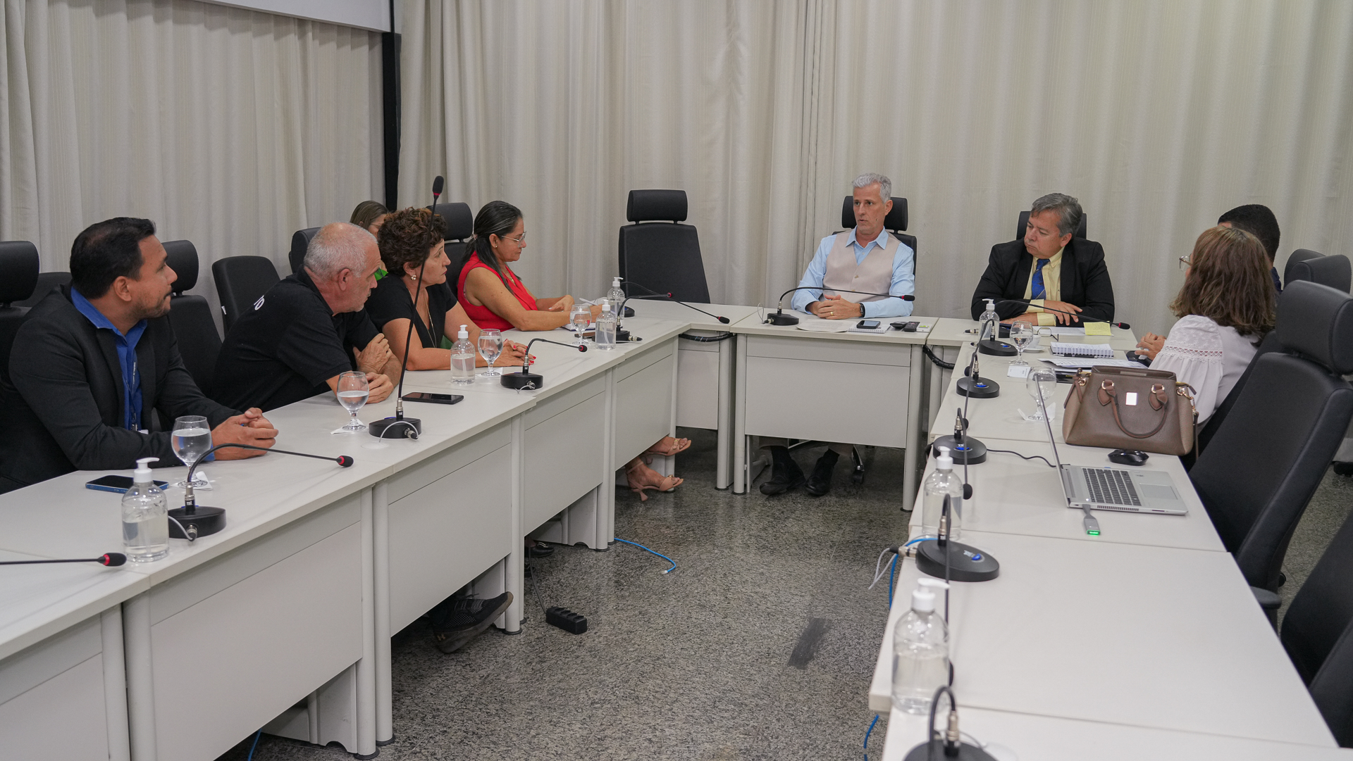 Em uma sala de reuniões com mesas brancas e cadeiras pretas, várias pessoas estão reunidas discutindo demanda dos servidores  