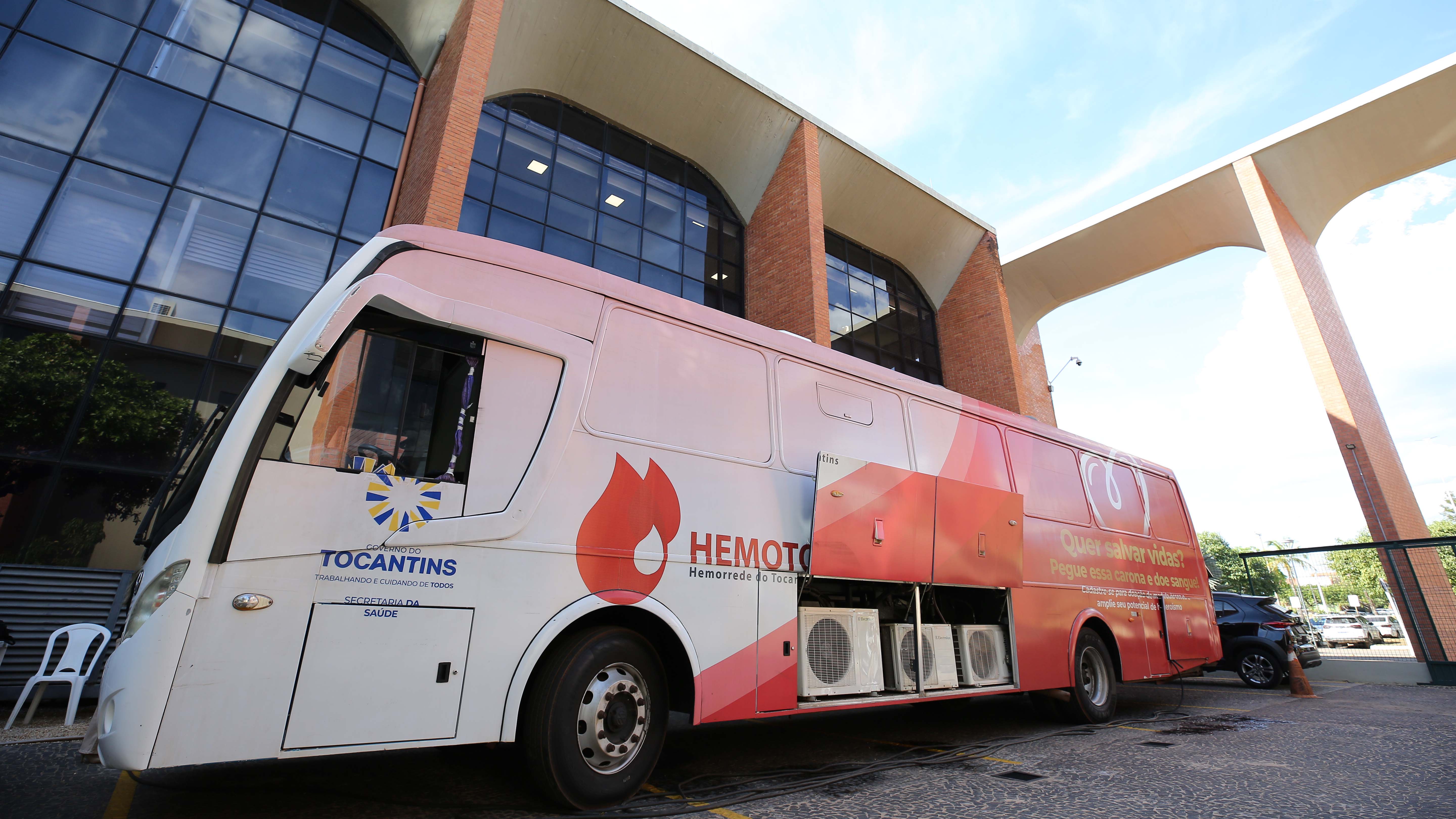 Imagem mostra o ônibus do Hemocentro estacionado no estacionamento do Tribunal de Justiça, ao fundo parte do prédio do Tribunal.