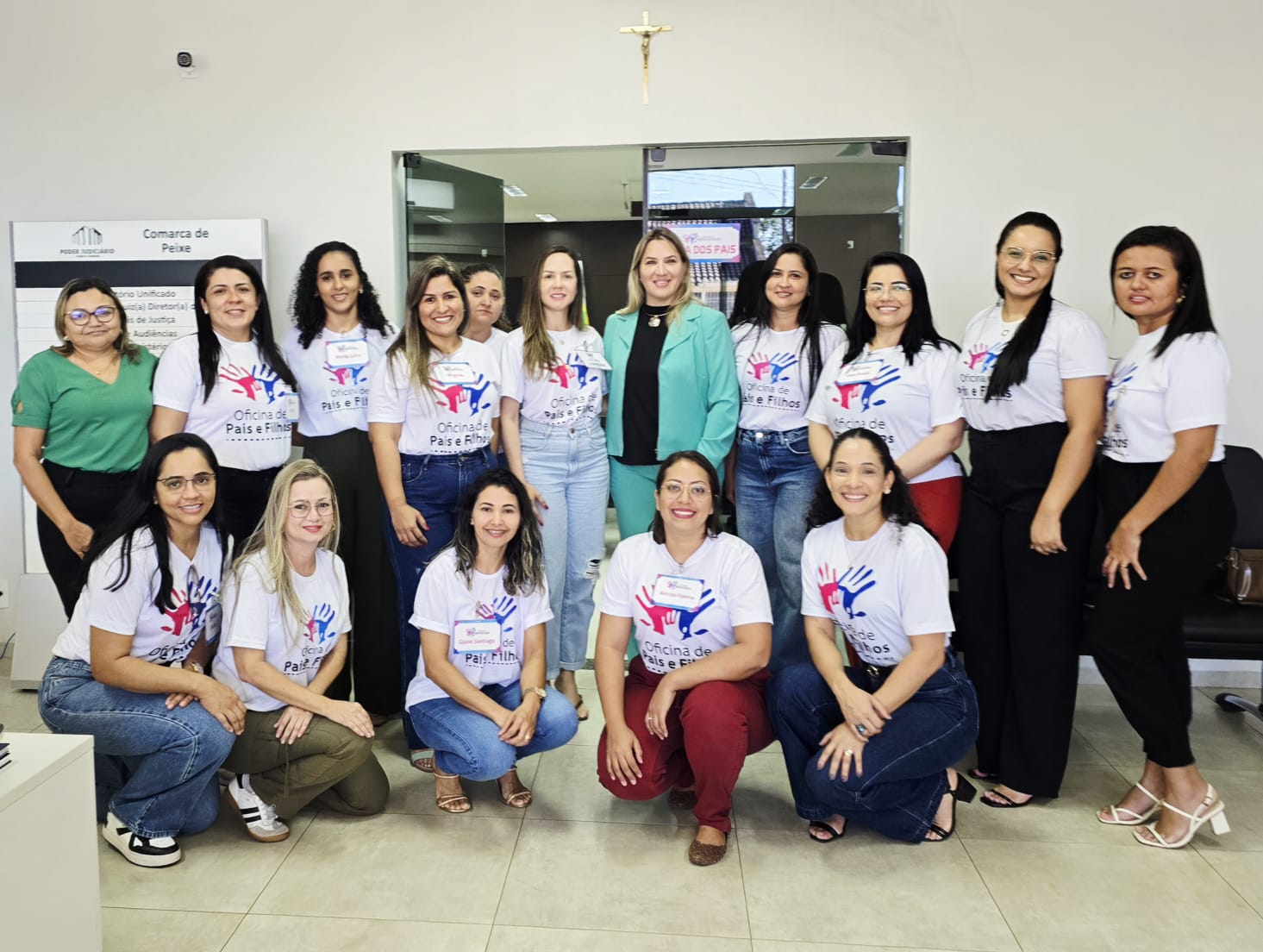 Dezesseis mulheres posam para foto; 11 estão em pé e cinco agachadas; a maioria usa camisetas brancas com logo e nome: Oficina de Pais e Filho