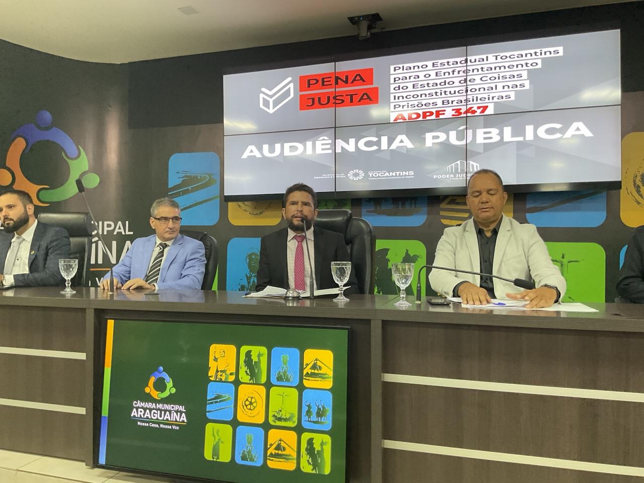 Imagem de uma tribuna com quatro autpridades sentadas, ao centro, de terno escuro, camisa branca e gravata vermelha, o juiz Eustáquio fala ao microfone; ao fundo um banner da audiência pública Pena Justa