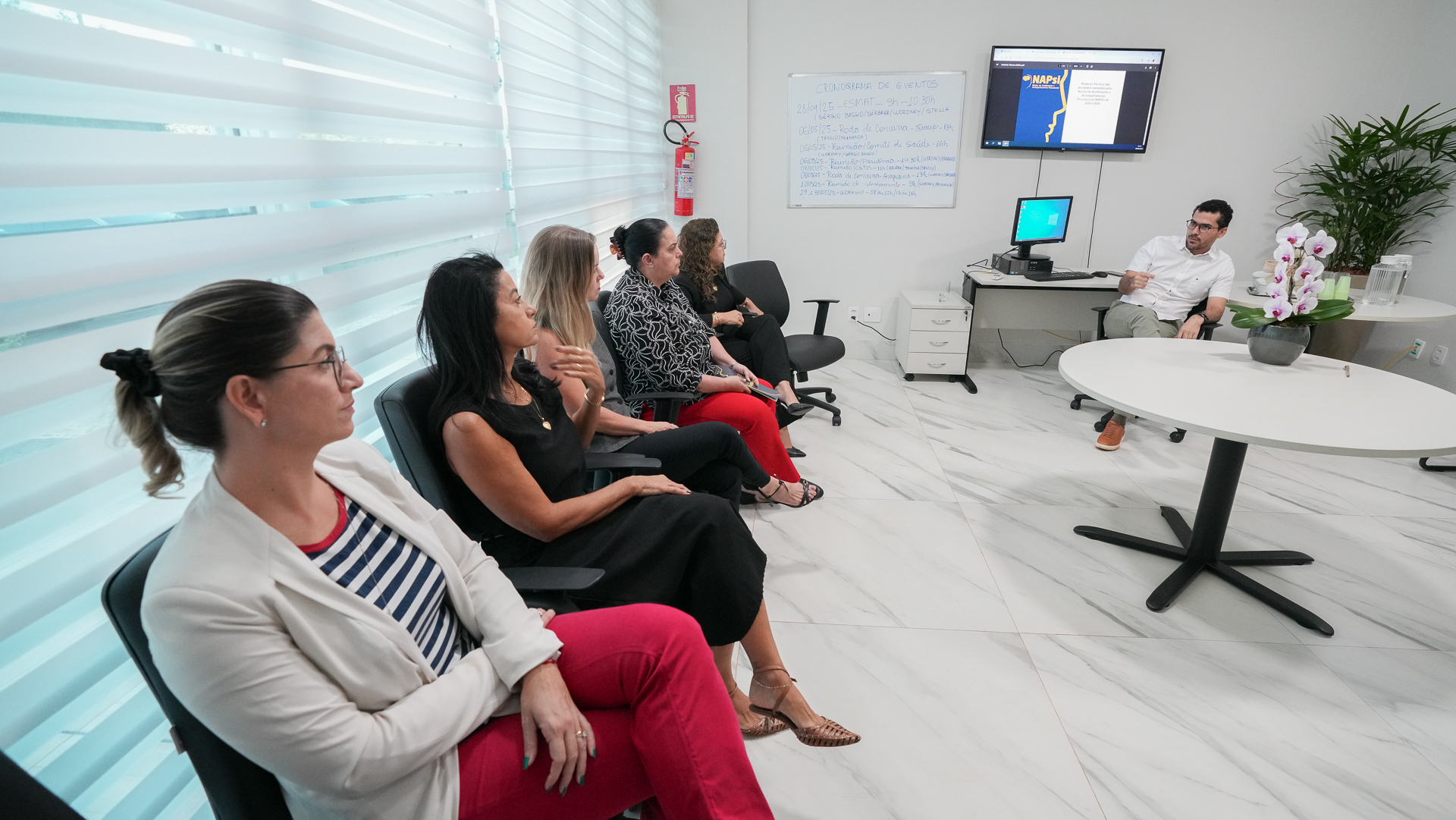 A imagem mostra um grupo de pessoas sentadas em uma disposição semi-circular em torno de uma mesa redonda, em uma sala.  