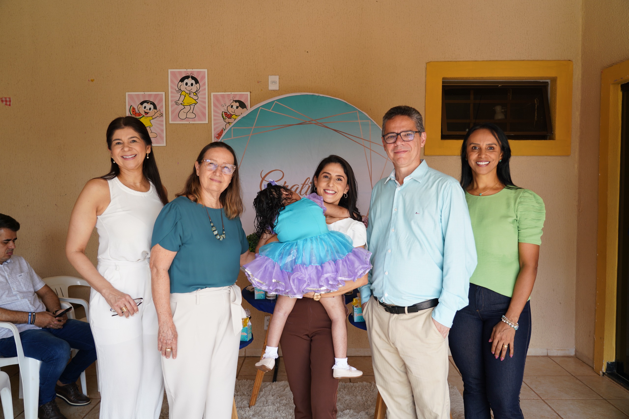 Imagem mostra cinco pessoas adultas, sendo uma mulher ao centro segurando uma criança com roupa nos tons de azul e lilás; todos estão em pé e posando para a foto; à direita dela está o juiz Adriano e à esquerda, a desembargadora Maysa.