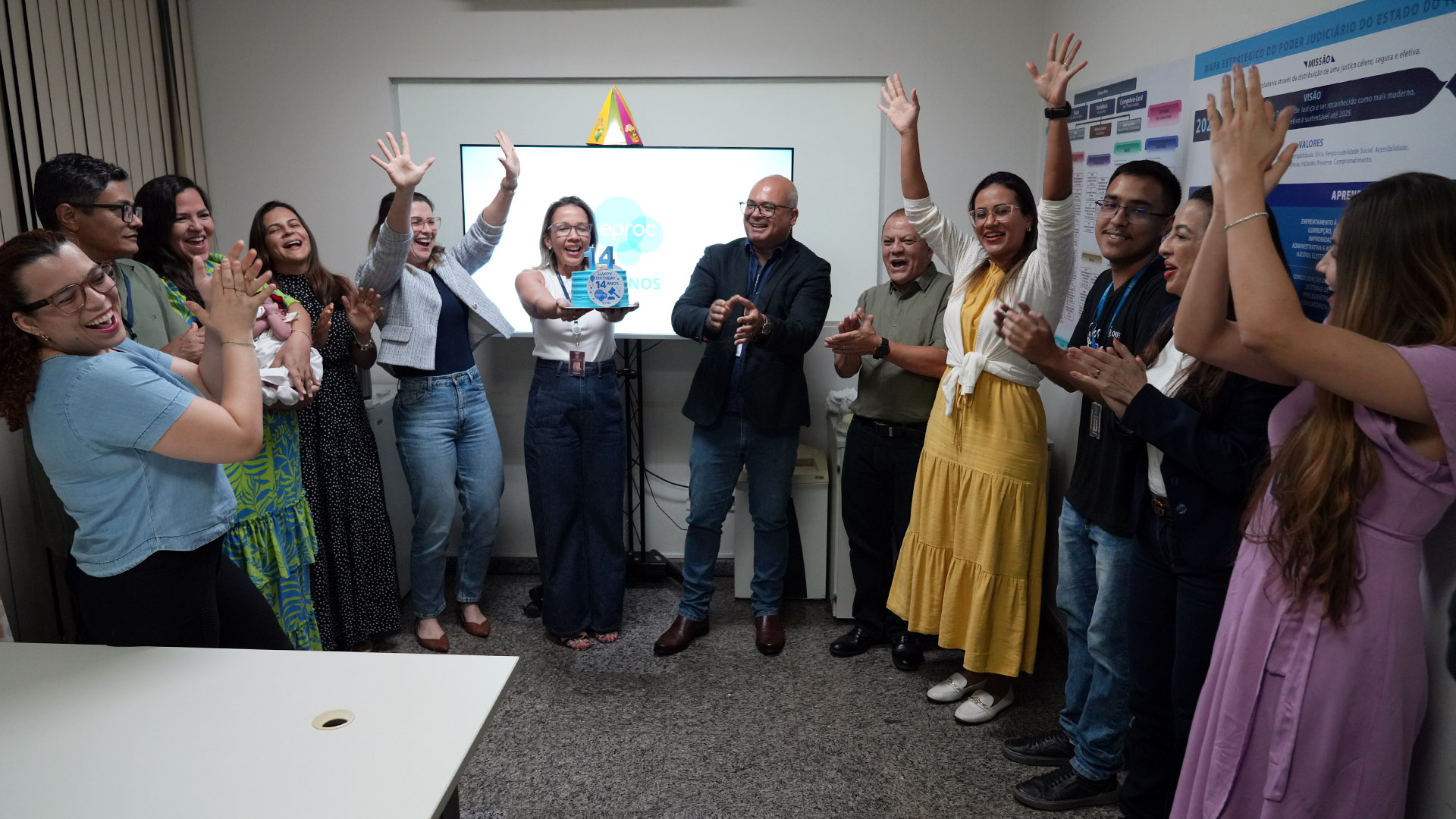 A imagem mostra um grupo de pessoas reunido para celebrar os 14 anos do sistema eproc. No centro, uma pessoa segura um bolo de aniversário, enquanto os demais participantes demonstram entusiasmo — alguns sorriem e outros aplaudem. A cena transmite um momento de comemoração institucional, com clima descontraído e participativo.