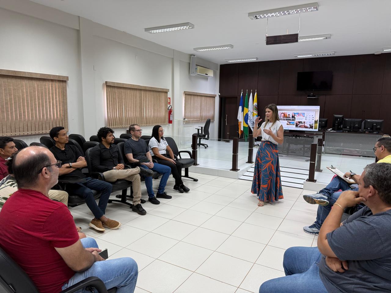 A imagem mostra um grupo de pessoas em uma sala de reuniões. No centro, uma mulher está em pé, falando para um grupo que está sentado em cadeiras organizadas em formato circular. As pessoas estão atentas.