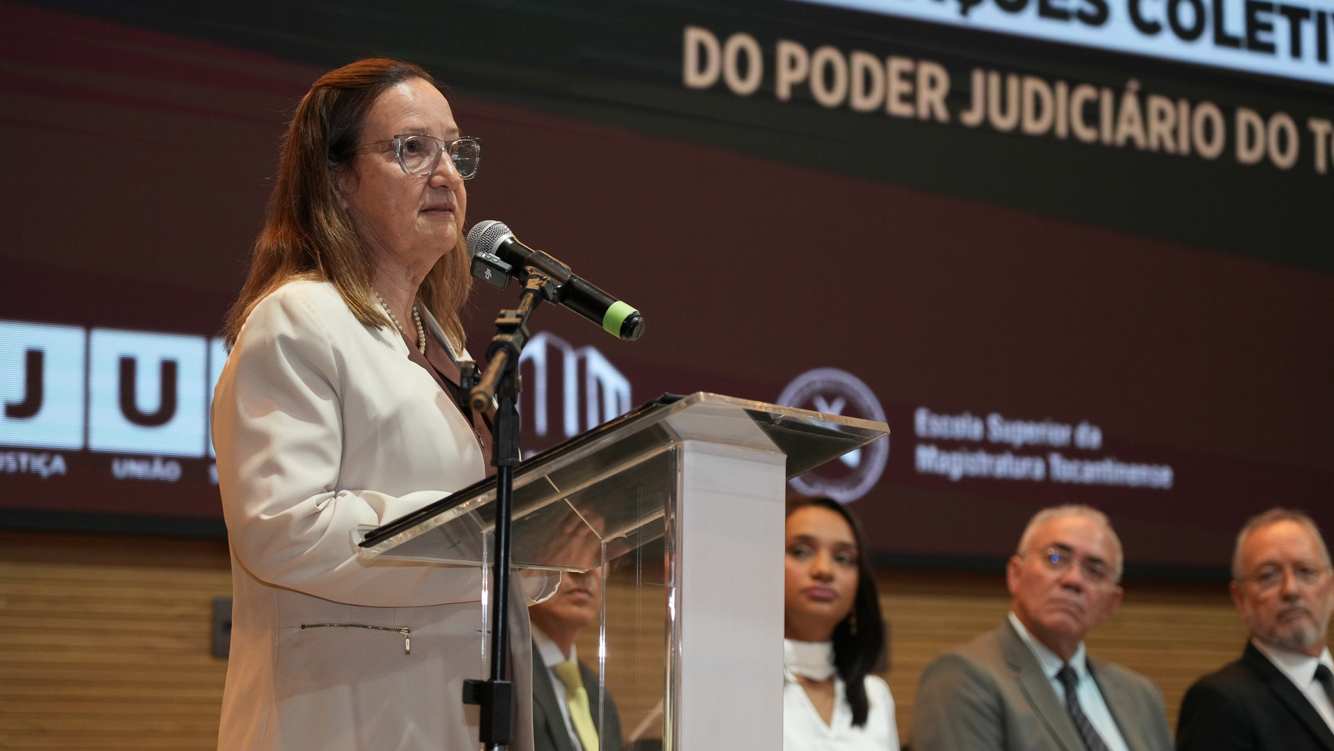 Presidente do TJTO fala ao microfone, uma mulger de pele clara, usa óculos e roupa marrom e blazer claro
