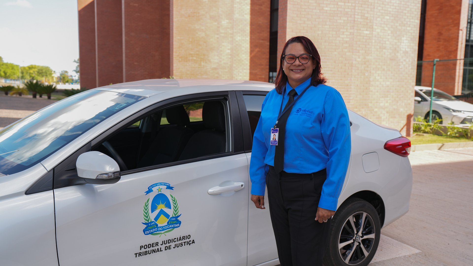 A imagem mostra uma mulher sorridente em pé ao lado de um carro branco institucional do Tribunal de Justiça do Estado do Tocantins (TJTO). Ela veste uniforme azul com gravata preta, crachá de identificação e óculos. Ao fundo, vê-se parte do prédio do TJTO, com fachada de tijolos e um ambiente externo bem cuidado, com plantas e calçamento. A cena transmite profissionalismo e acolhimento.