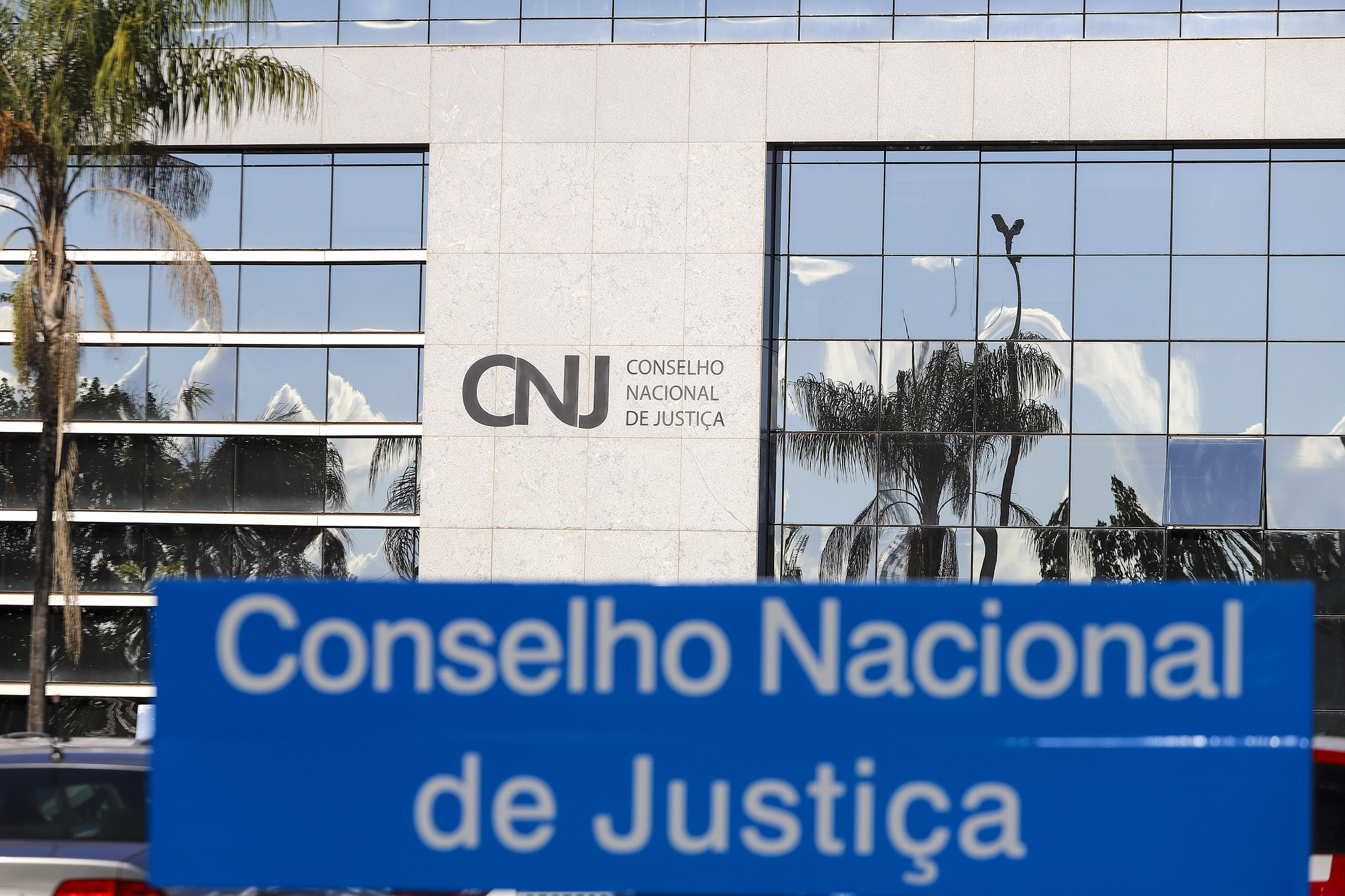 Foto da fachada da sede do CNJ com a placa de identificação em azul em primeiro plano e uma palmeira ao fundo, à esquerda