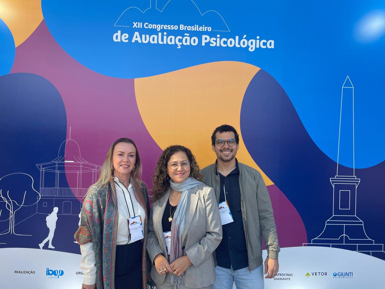 Descrição da imagem (da esquerda para a direita): Os psicólogos Janaína Araújo, Bárbara Camargo e Tássio Soares posam sorridentes em frente ao painel oficial do XII Congresso Brasileiro de Avaliação Psicológica. O fundo colorido em tons de azul, laranja e roxo traz o nome do evento em destaque, além de ilustrações estilizadas de monumentos e edificações. Todos vestem roupas sociais e usam crachás de identificação do congresso. O clima parece ameno, sugerindo temperaturas mais baixas.