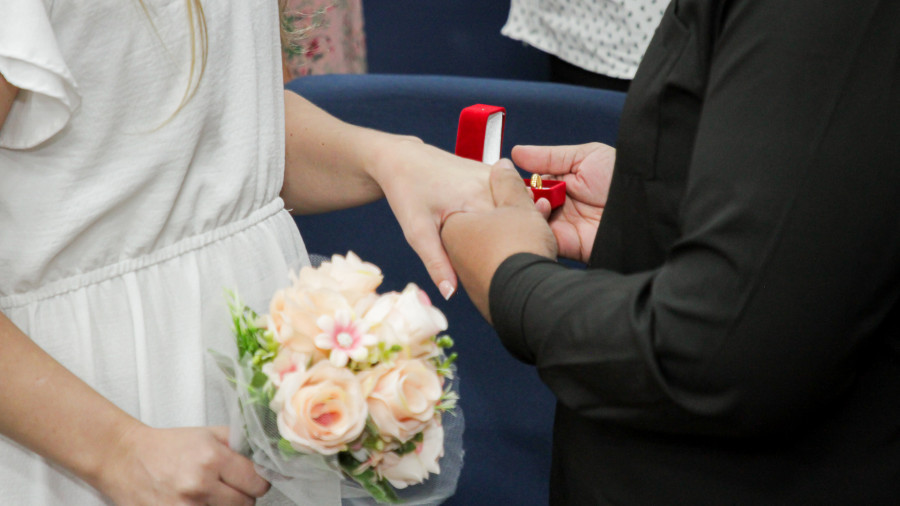 imagem de detalhe das mãos de um casal durante troca de alianças; na mão esquerda da noiva, ela segura um bouquet de flores rosas 