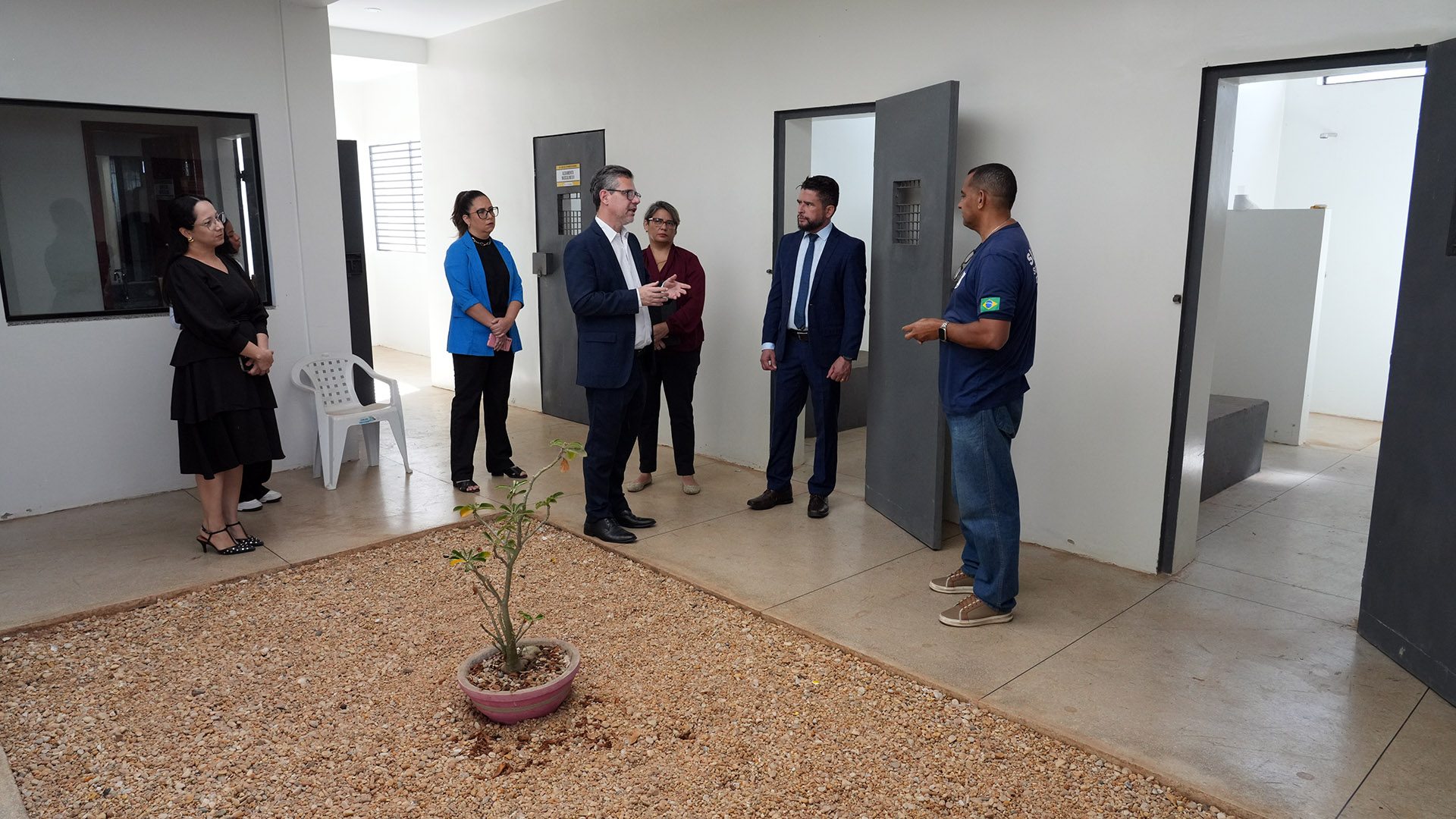 A imagem mostra um grupo de sete pessoas em visita a um espaço institucional.  No centro, um homem de terno azul claro e óculos conversa com os demais, gesticulando com as mãos. Ao lado dele, outro homem de terno azul escuro e gravata observa com atenção, enquanto um servidor, vestido com camiseta azul com a bandeira do Brasil na manga, parece explicar detalhes sobre o local.  Atrás deles, três mulheres acompanham a visita: uma com blazer azul, outra com blusa vinho e a terceira vestida de preto. Mais ao fundo, é possível ver portas de ferro abertas, que lembram celas ou salas de segurança.