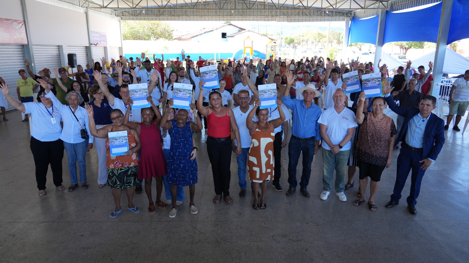 A imagem mostra um grande grupo de pessoas organizadas em fileiras, ocupando quase toda a extensão horizontal do espaço. Elas estão em pé durante a 3ª Semana Nacional de Regularização Fundiária nas Serras Gerais. Algumas seguram seus títulos de imóveis levantados, em gesto de comemoração pela conquista.
