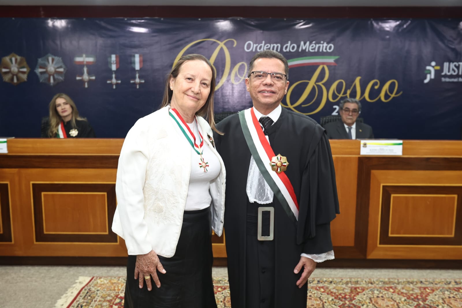 Duas pessoas posam lado a lado em uma cerimônia de condecoração da Ordem do Mérito Dom Bosco. À esquerda, a presidente do TJTO veste blusa branca e blazer claro, usando no peito a medalha da Ordem. À direita, o desembargador do TRT-10 José Ribamar  trajando toga preta com faixa verde, branca e vermelha, também usando a medalha no peito. Ao fundo, mesa de autoridades com duas pessoas sentadas e um grande painel azul com o título “Ordem do Mérito Dom Bosco” em dourado