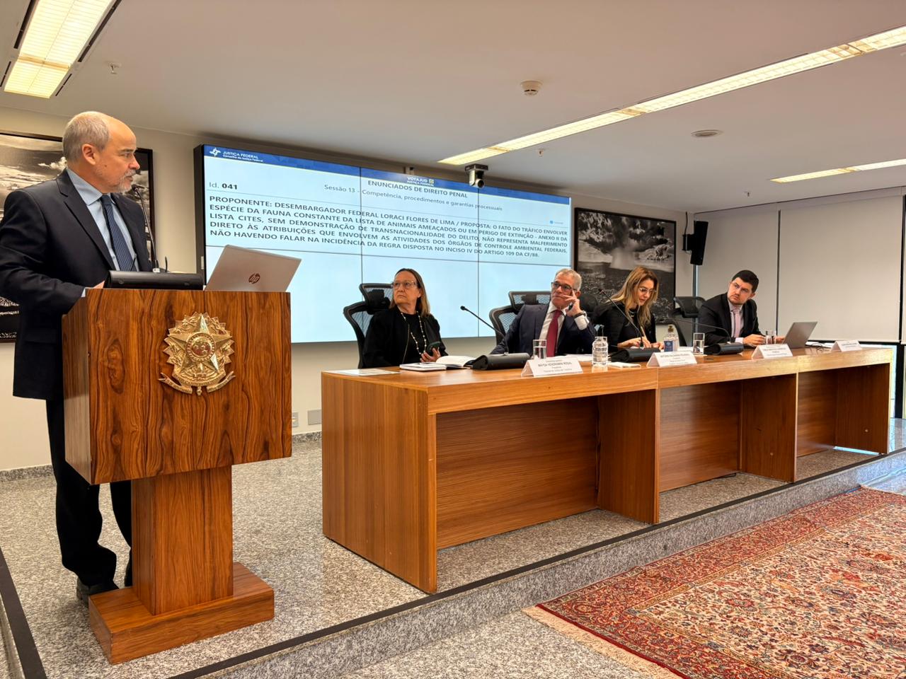 Imagem de um evento em uma sala, com pessoas sentadas em torno de uma mesa e um orador em um púlpito
