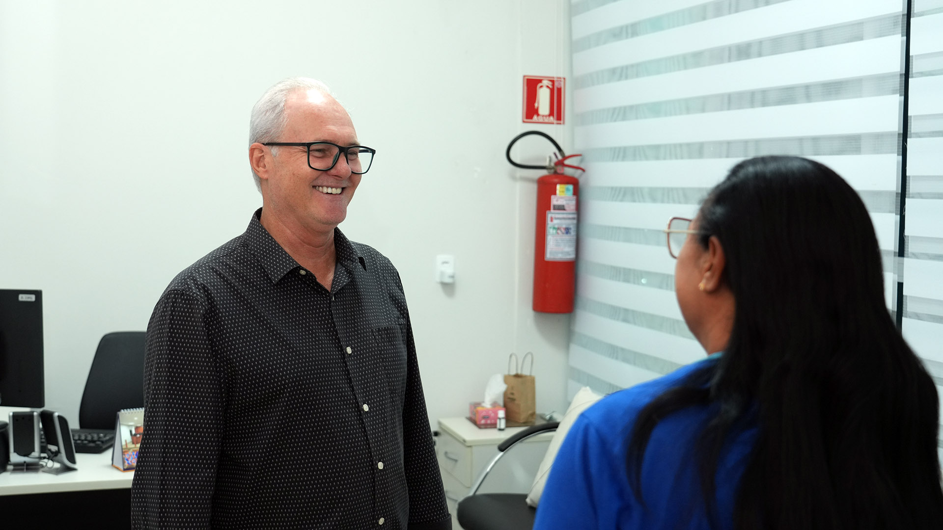 Psicólogo do NAPsi, homem de cabelos brancos, usando óculos e camisa cinza, sorrir para uma mulher que está de costa e de frente para ele; ela usa camiseta azul. 