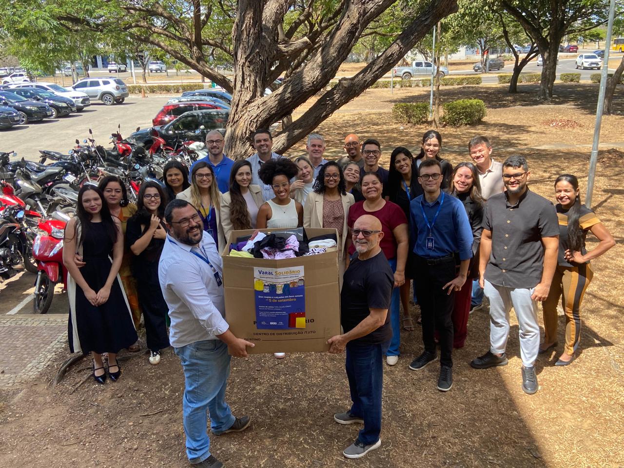 Em um local aberto, com árvores ao fundo, um grupo de pesspas posam pra foto com uma caixa cheia de  roupas para doação; dois homens estão à frente segurando a caixa.
