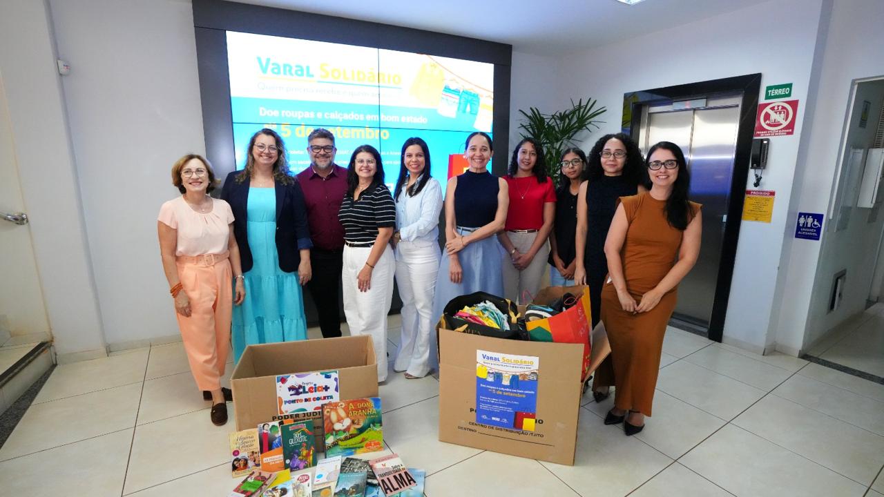 Dez pessoas aparecem em pé, à frente de um telão e atrás de duas caixas grandes com roupas doadas