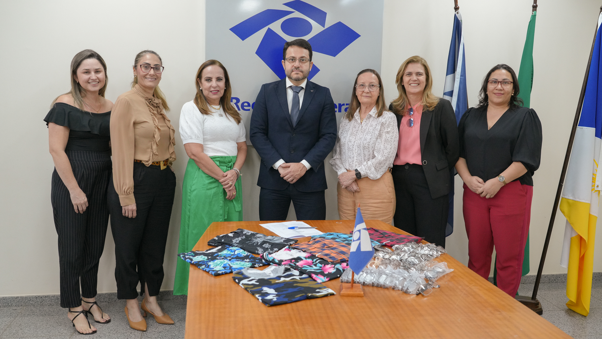 Sete pessoas em pé, de frente, sendo seis mulheres e um homem, posam para foto, ao fundo a logo da Receita Federal e à frente uma mesa com algumas peças de roupa