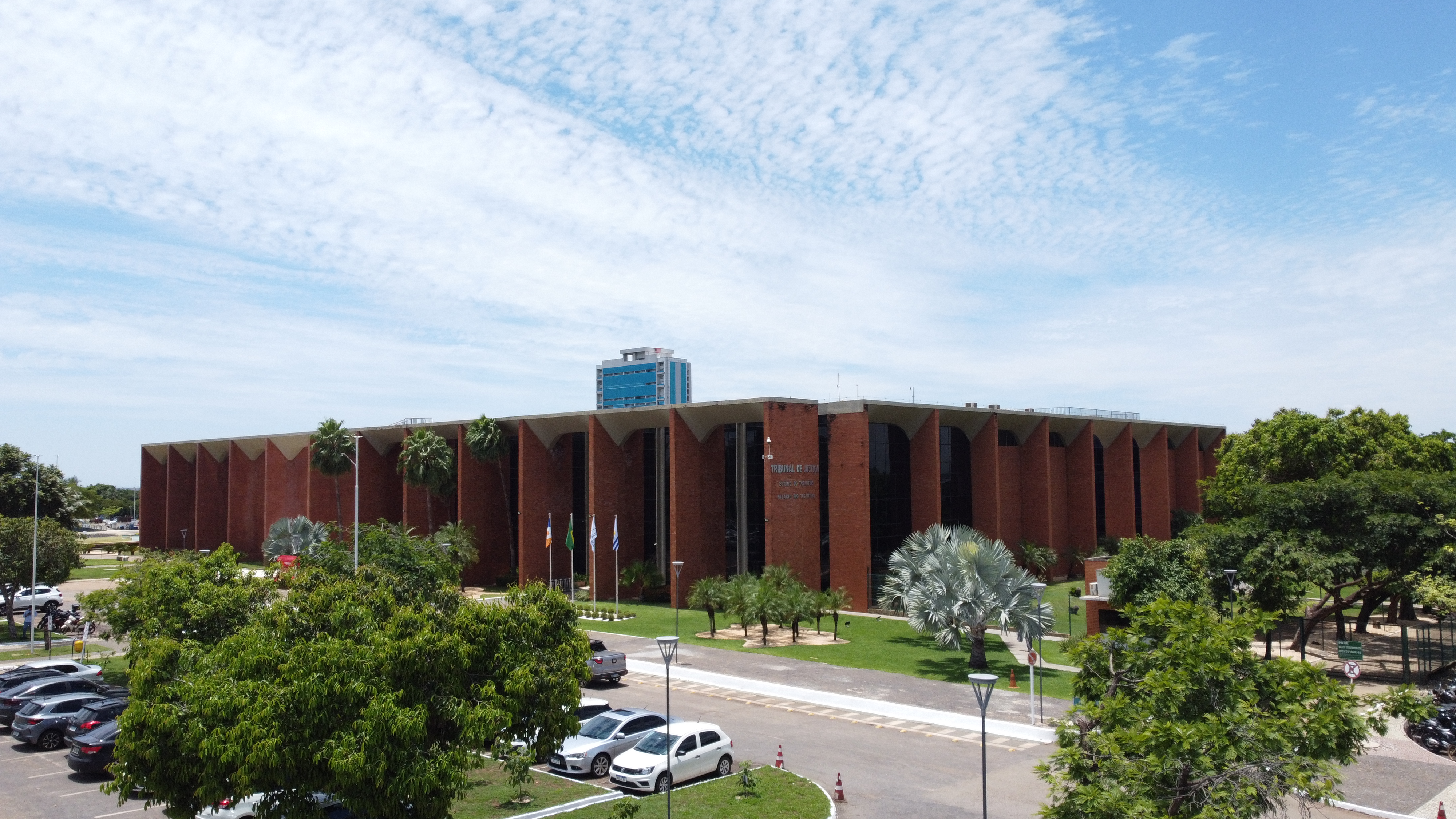 Na imagem aparece a fachada do prédio do Tribunal de Justiça do Tocantins, em Palmas, cercado por árvores, bandeiras e céu azul com nuvens claras.