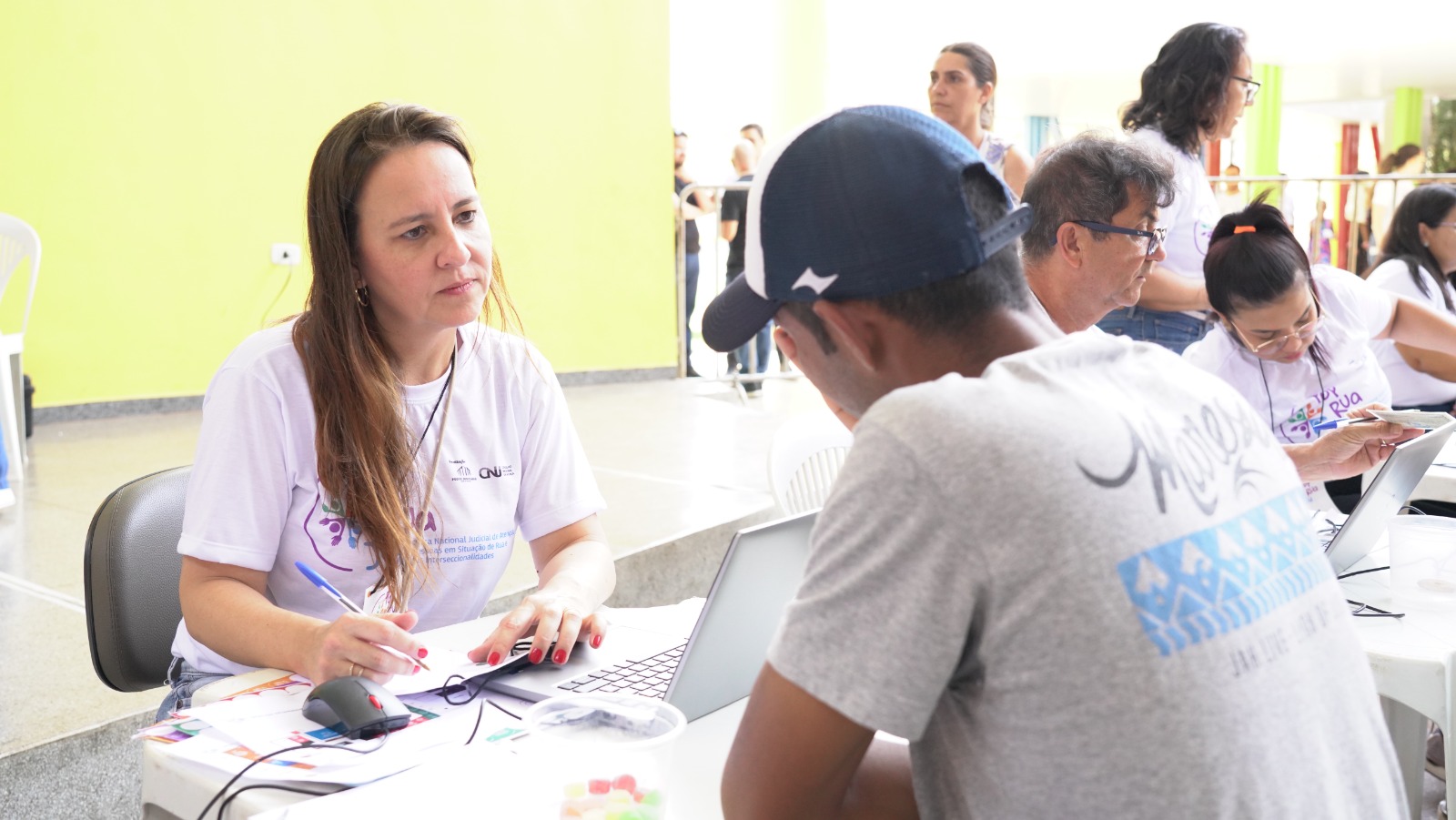 Na imagem aparece uma mulher de camiseta branca atende homem de boné azul em mesa com notebook e papéis. Ao fundo, outros voluntários prestam atendimentos no Pop Rua Jud.