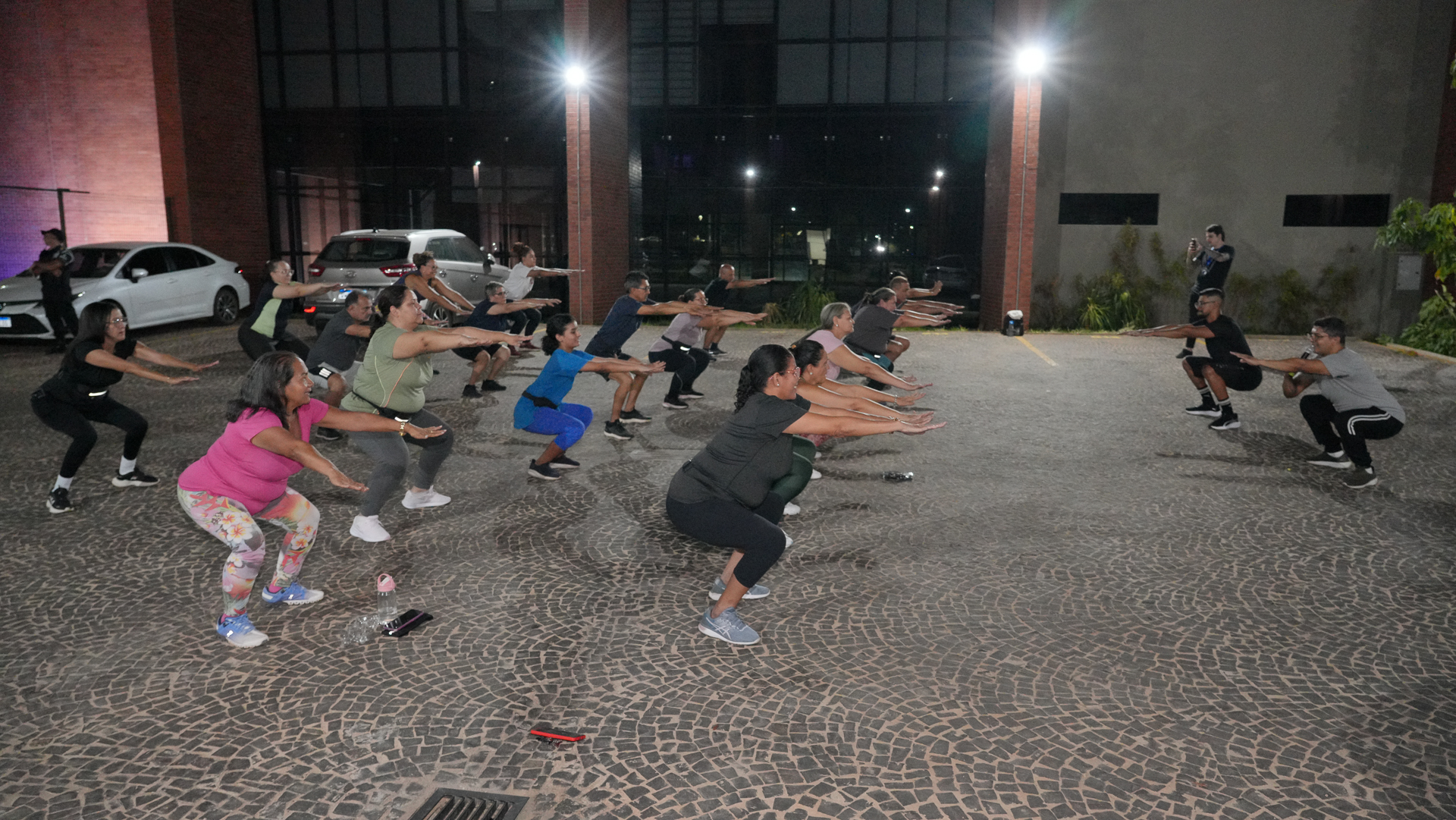 A foto mostra um grupo de pessoas participando de uma atividade física coletiva ao ar livre, provavelmente em frente ao prédio do TJTO. Elas estão alinhadas em fileiras, executando o movimento de agachamento, com os braços estendidos à frente. O ambiente é noturno, iluminado por refletores, e o piso é de pedras portuguesas.
