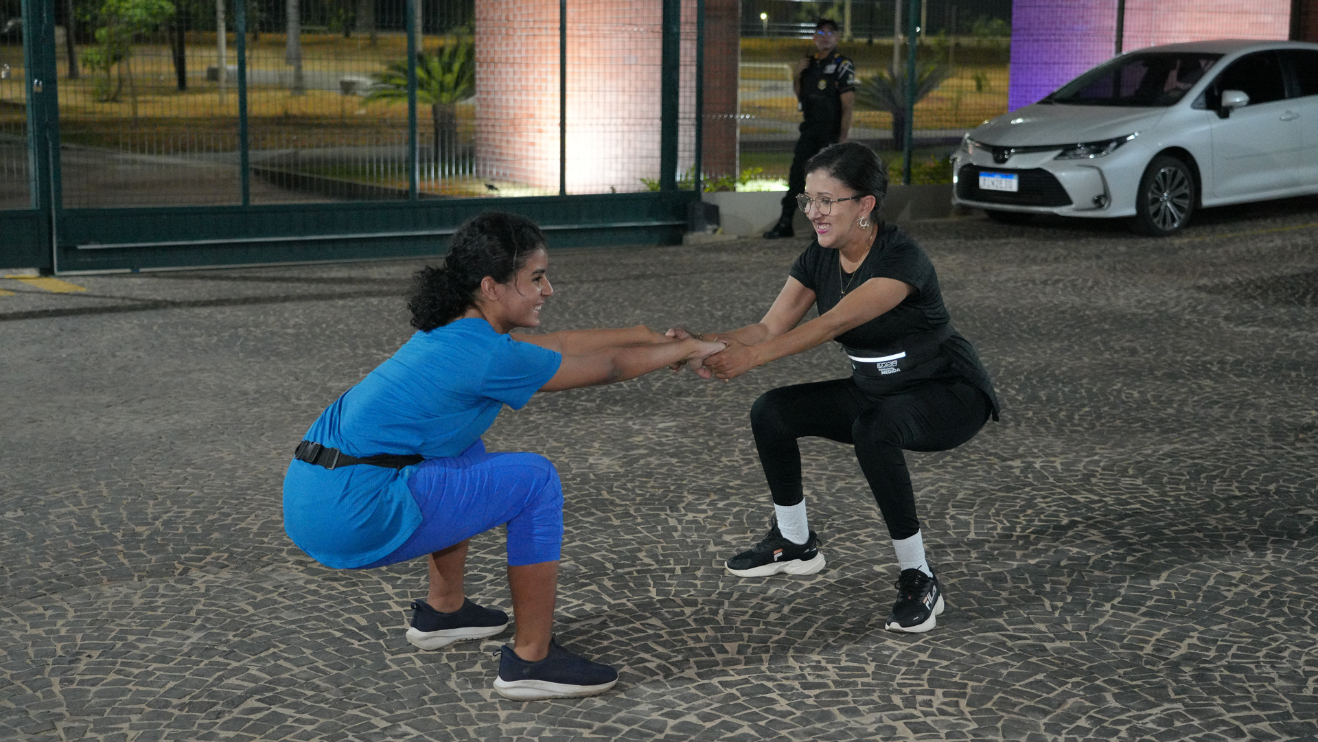 A foto mostra duas mulheres realizando um exercício físico em dupla, ao ar livre, sobre um piso de pedras.