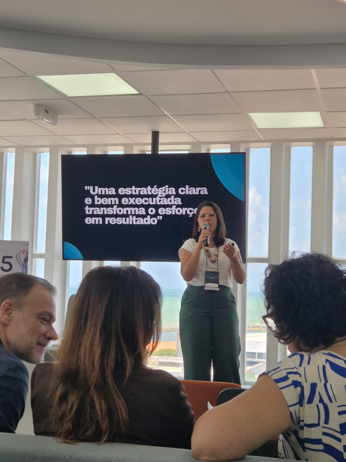 Servidora do TJTO ministra palestra, em pé; ela fala ao microfone e ao fundo um painel com fundo azul com os seguintes dizeres: "Uma estratégia clara e bem executada transforma o esforço em resultado" 