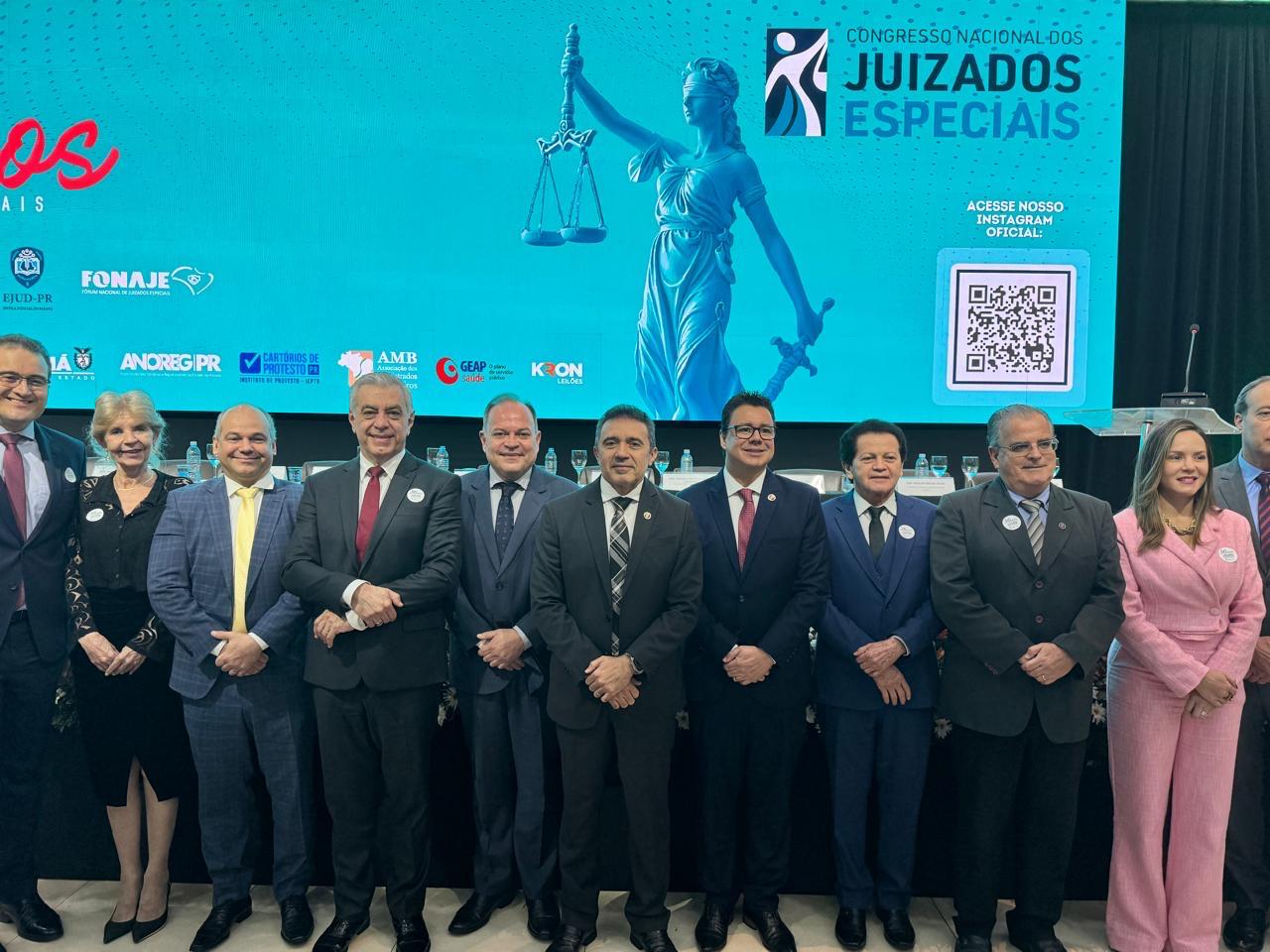A imagem traz um grupo de 11 pessoas posa lado a lado, sorrindo, em frente a um grande painel digital azul com a inscrição “Congresso Nacional dos Juizados Especiais”. O painel exibe a imagem de uma estátua estilizada da deusa da Justiça segurando a balança, além de logomarcas de instituições apoiadoras e um QR Code.  As pessoas estão em trajes formais — ternos e vestidos sociais. A maioria dos homens veste terno escuro com gravata, enquanto uma das mulheres, à direita, usa um conjunto social rosa claro. Outra mulher, mais à esquerda, veste preto com detalhes em branco.