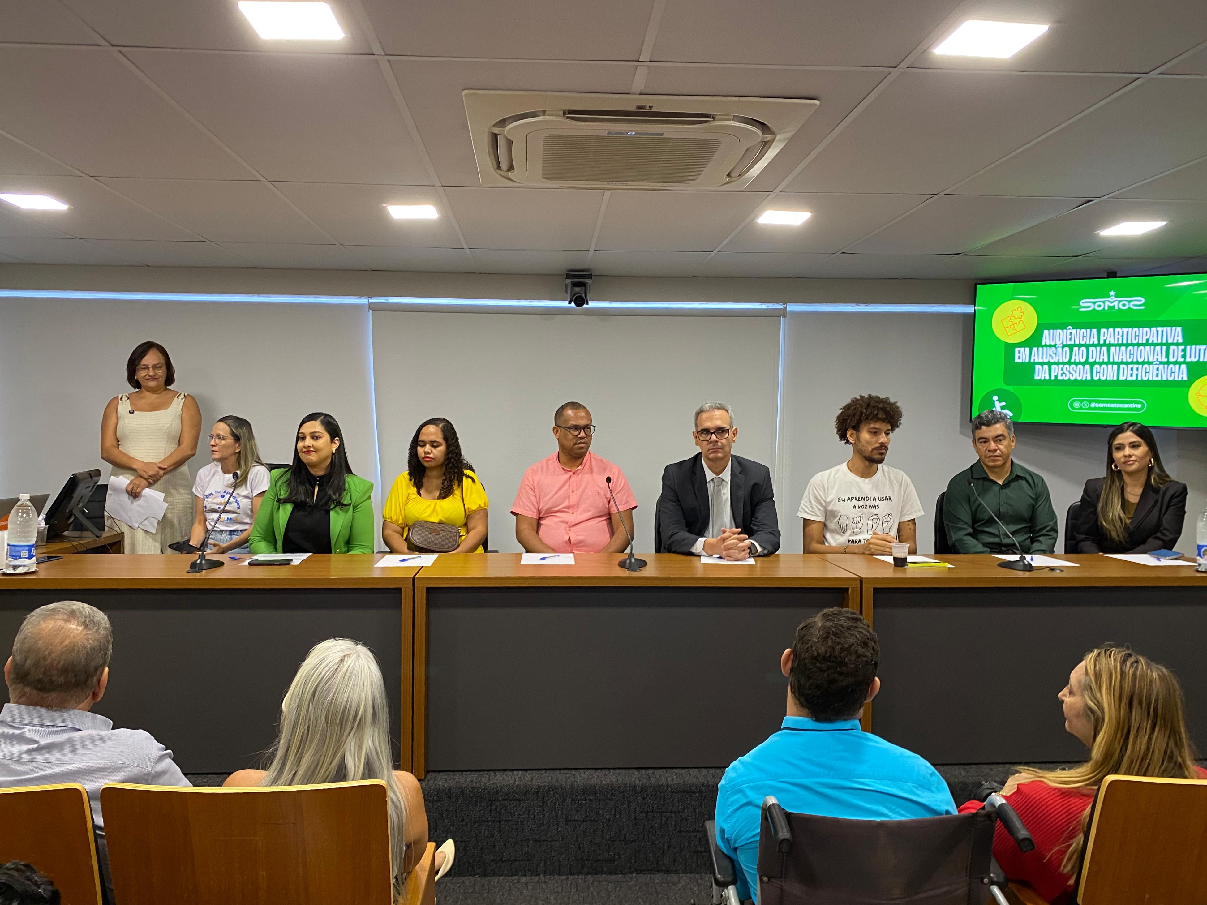 A fotografia mostra uma mesa de autoridades dispostas em auditório, na Câmara Municipal de Palmas. O ambiente tem iluminação clara, teto branco e uma tela verde ao fundo com os dizeres: “Audiência Participativa em alusão ao Dia Nacional de Luta da Pessoa com Deficiência – Somos”.  Sentados à mesa, da esquerda para a direita, estão: uma mulher de blazer verde, uma mulher de blusa amarela, um homem de camisa rosa, um homem de terno preto, um homem de camiseta branca, um homem de camisa verde e uma mulher de blazer preto.  Ao lado esquerdo, em pé, há duas mulheres: uma veste vestido claro e segura folhas nas mãos, e a outra usa camiseta branca estampada.