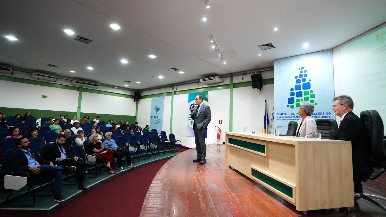 A imagem mostra o desembargador Marco Villas Boas falando ao microfone durante a conferência “Inteligência Artificial na Gestão Pública”, realizada na VI Semana Acadêmica de Gestão Pública do IFTO – Câmpus Palmas. Ele está diante de uma plateia em um auditório, enquanto duas pessoas permanecem sentadas à mesa ao seu lado.