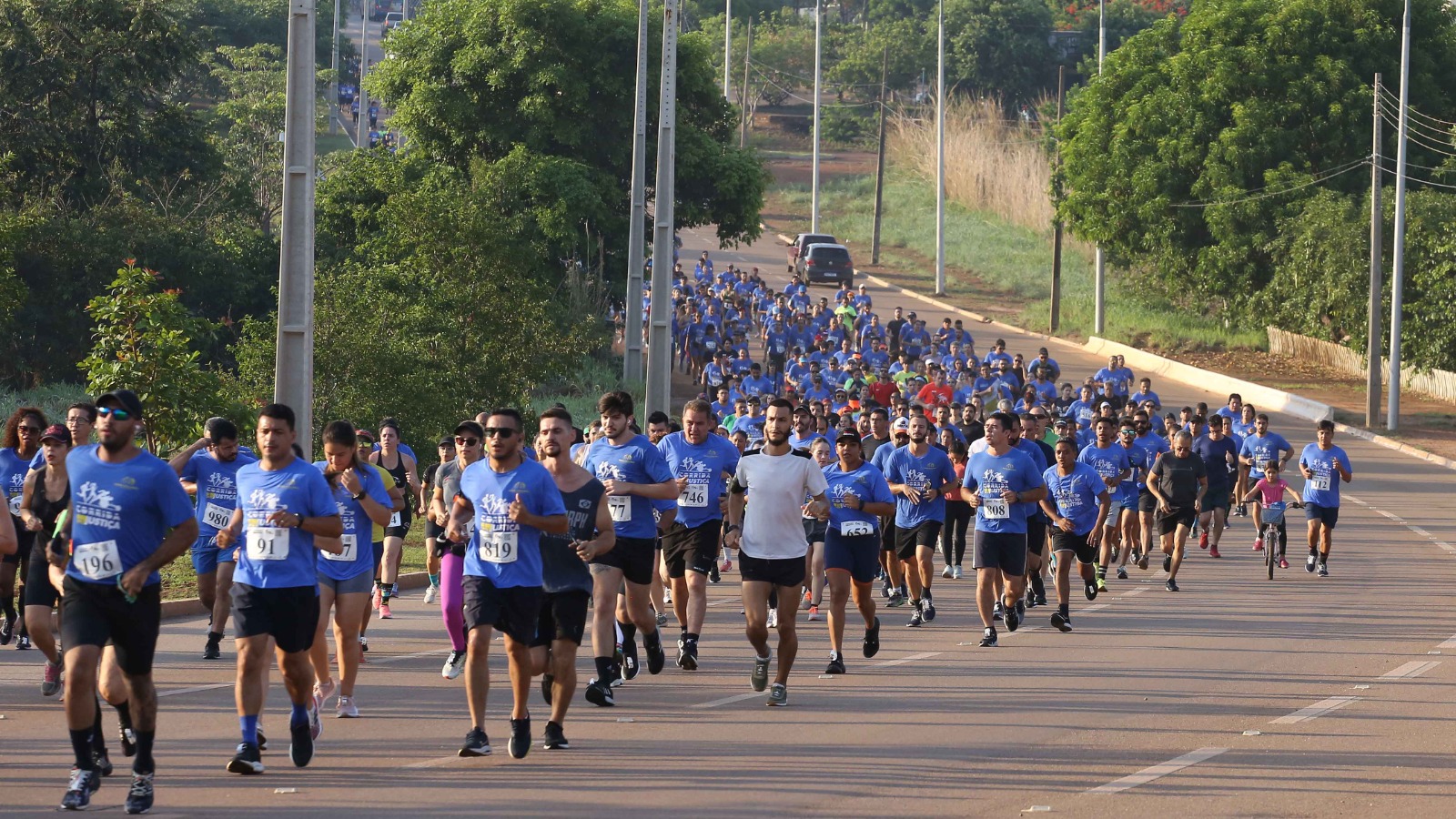 A foto mostra uma grande corrida de rua em uma avenida larga, durante o dia. Centenas de pessoas participam do evento esportivo, a maioria vestindo camisetas azuis e números de identificação presos à frente da roupa. Os corredores ocupam quase toda a via, que está cercada por árvores e postes de iluminação. Em primeiro plano, vê-se alguns atletas já mais próximos da câmera, correndo em diferentes ritmos; no fundo, uma multidão se estende até o horizonte, dando a sensação de movimento e energia coletiva. À direita, uma criança aparece pedalando uma bicicleta, acompanhando os participantes. O clima é de união, esforço físico e celebração esportiva.