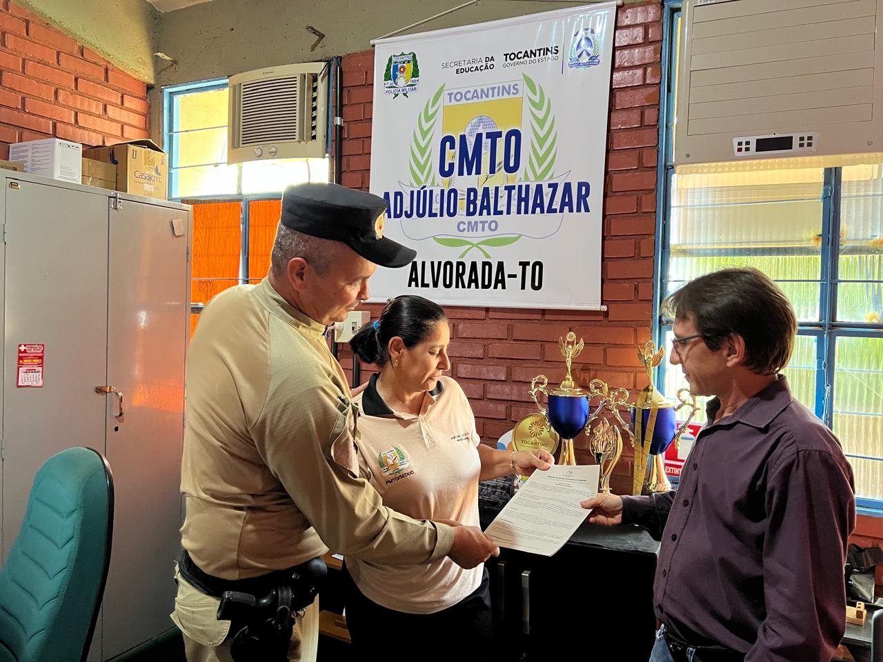 A imagem mostra três pessoas observando um documento com informações sobre o Concurso de Criação da Logomarca do Projeto Alvorada Sustentável, durante o momento de divulgação da iniciativa.