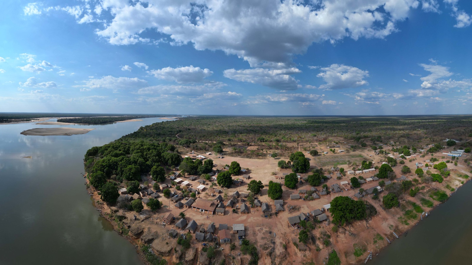 A imagem aérea mostra uma aldeia localizada na confluência de dois rios, cercada por uma vasta paisagem natural, sob um céu azul vibrante.