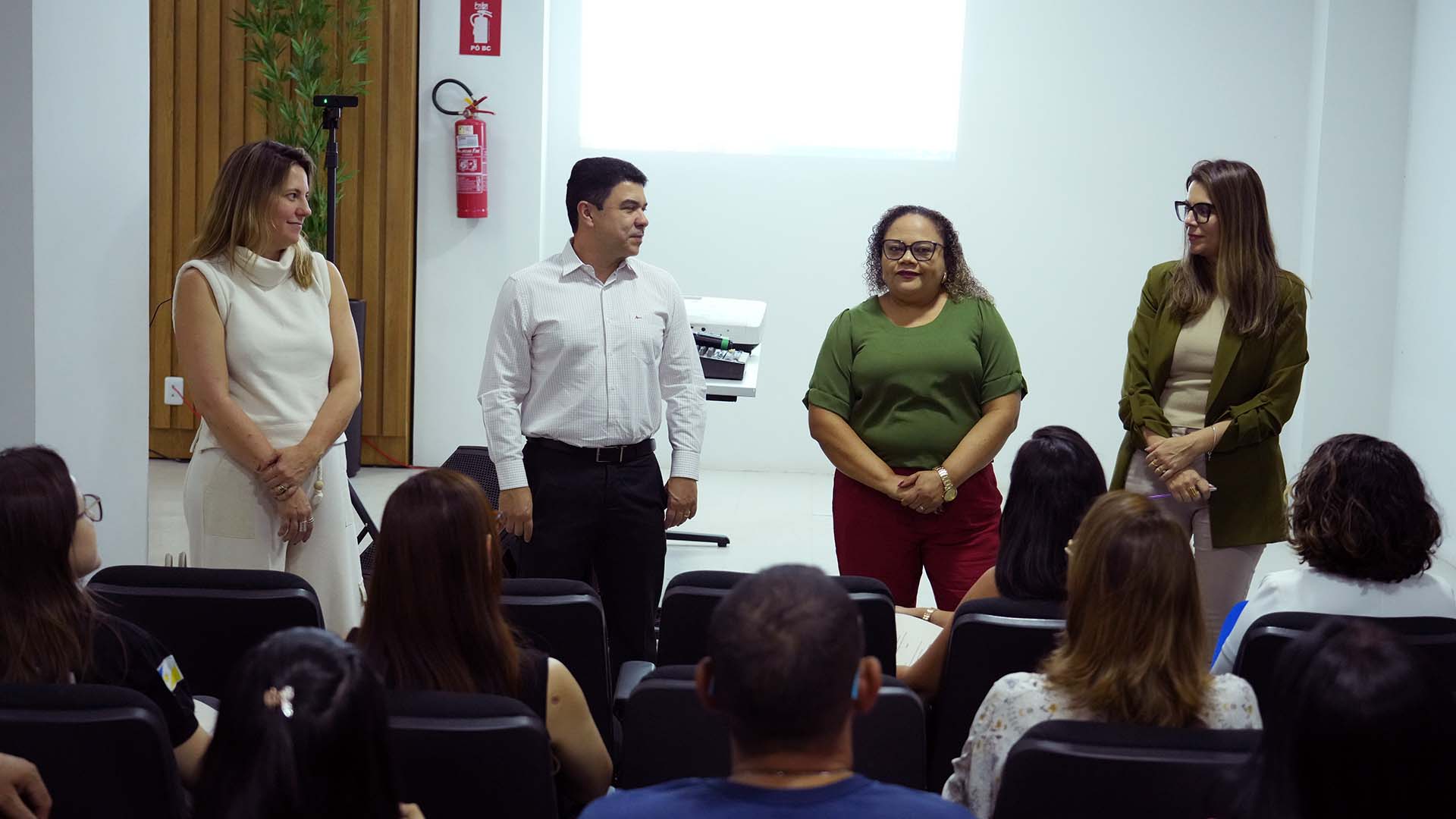 A imagem mostra um auditório com pessoas sentadas assistindo a quatro palestrantes em pé à frente. Da esquerda para a direita: uma mulher de roupa clara sem mangas; um homem de camisa branca e calça preta; uma mulher de blusa verde e calça vermelha; e outra mulher de blazer verde e calça clara. Ao fundo, há um projetor ligado, uma planta e um extintor na parede branca.