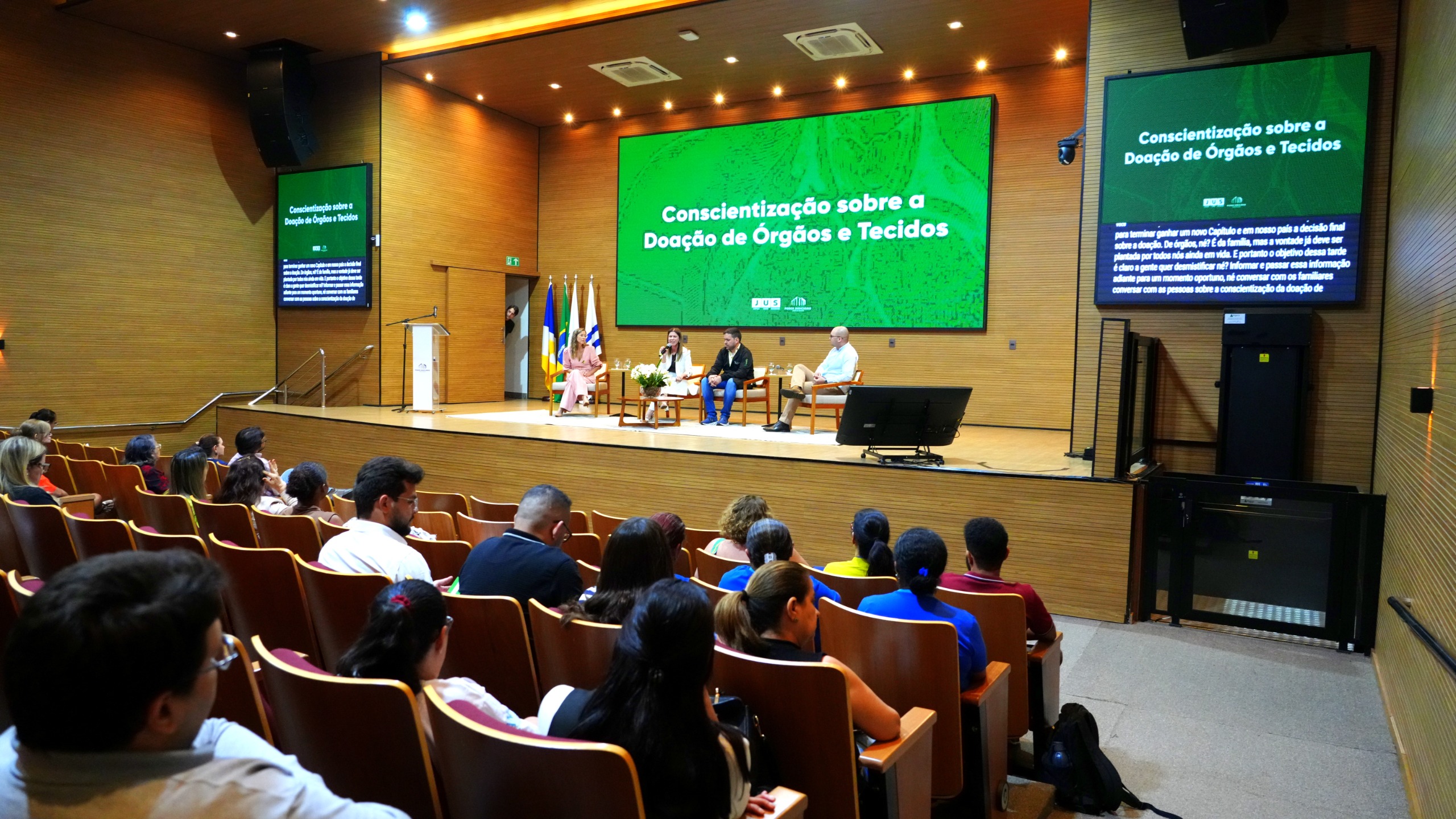 As imagens são do auditório do TJTO com paredes de madeira clara, palco com quatro cadeiras, mesa com flores e púlpito lateral. No fundo, duas telas verdes exibem “Conscientização sobre a Doação de Órgãos e Tecidos”. Três palestrantes e uma mediadora estão sentados no palco; ao lado, bandeiras do Brasil, Tocantins e TJTO. O público ocupa as primeiras fileiras em poltronas de madeira. Um telão lateral exibe legendas do evento, transmitindo ambiente formal e organizado.