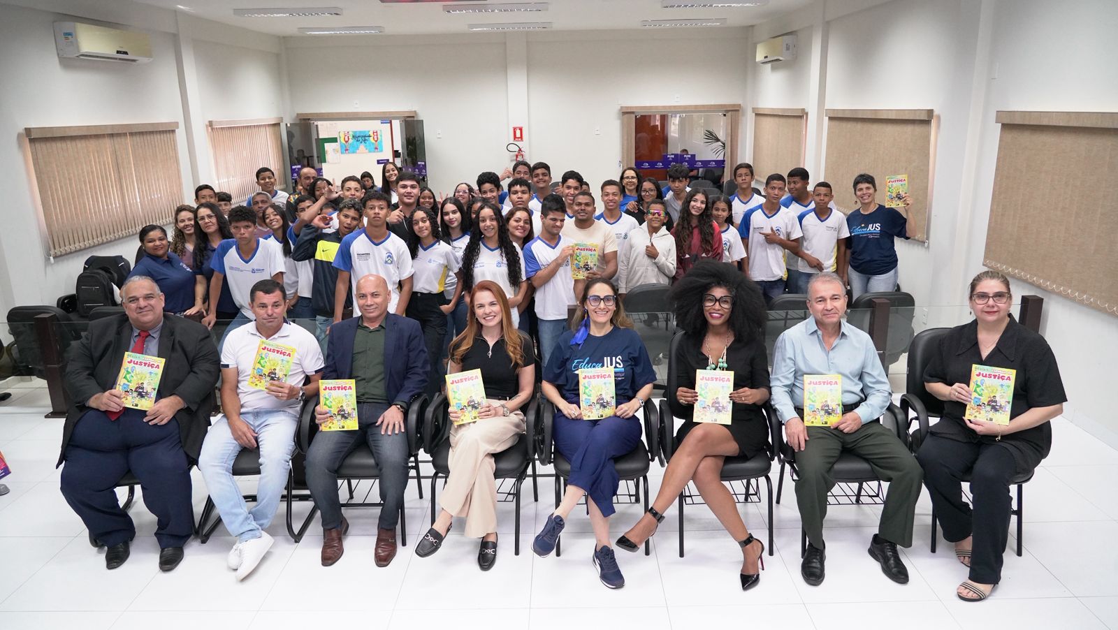 Homens e mulheres sentados em auditório em primeiro plano segurando uma cartilha de capa amarela. Atrás deles, estudantes em pé posando para foto