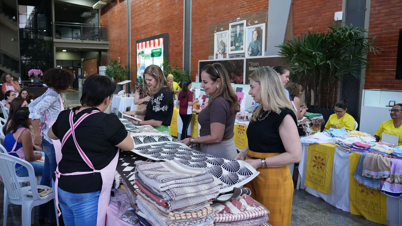 A imagem mostra a desembargadora Ângela Prudente e outras mulheres visitando a quarta edição da Feira Ecosolidária.com uma artesã em sua banca na Feira Ecosolidária. Ao fundo, outras bancas estão dispostas no hall do TJTO, com artesãs e visitantes circulando pelo espaço.