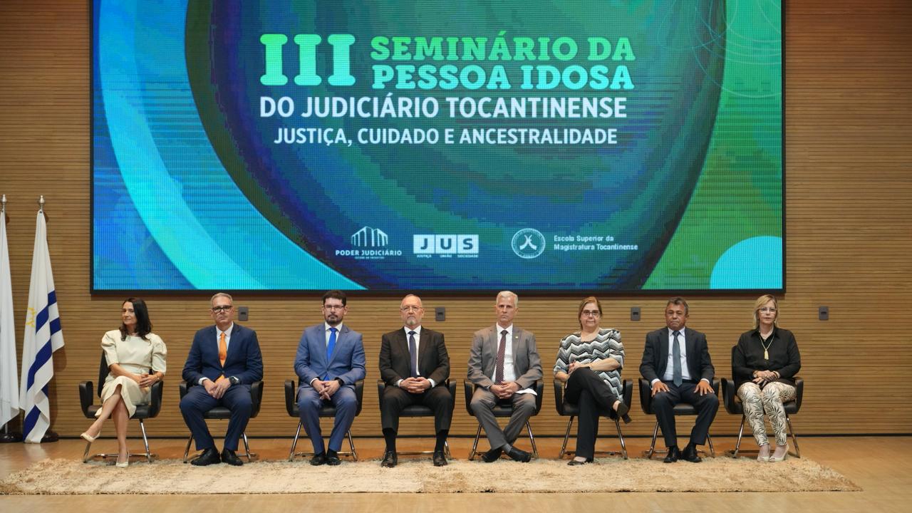 A imagem mostra oito pessoas sentadas em cadeiras dispostas em linha reta no palco. Ao fundo, um grande painel digital exibe o tema do evento: "III Seminário da Pessoa Idosa do Judiciário Tocantinense - Justiça, Cuidado e Ancestralidade". 