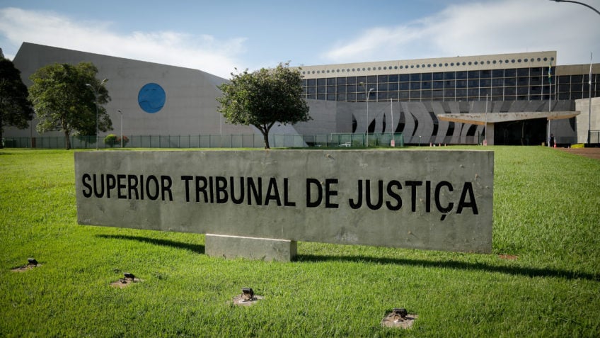 A imagem mostra a fachada do Superior Tribunal de Justiça em Brasília, com a placa institucional em primeiro plano e o prédio moderno ao fundo sob luz natural.