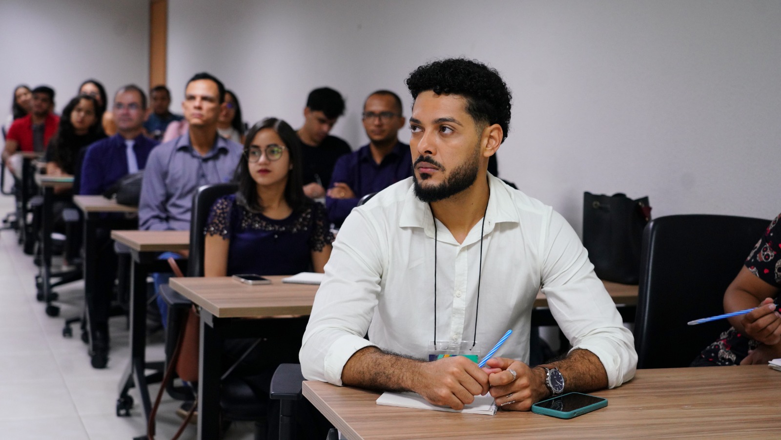 Imagem de servidores em sala de aula durante curso de capacitação. Em primeiro plano, homem pardo com cabelo cacheado aparado nas laterais e barba aparada, veste camisa branca