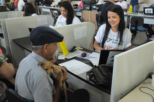 A imagem mostra um homem mais velho segurando um cachorro marrom enquanto é atendido por uma mulher sorridente atrás do balcão, com outra mulher atendendo uma pessoa fora do quadro, em um ambiente iluminado e descontraído.