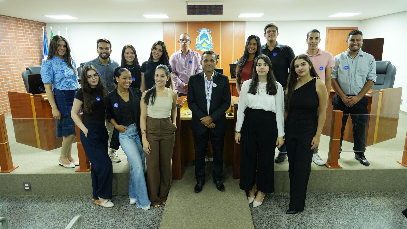 Homens e mulheres em pé posando pra foto dentro de sala de paredes brancas. Ao fundo, brasão do poder judiciário do Tocantins