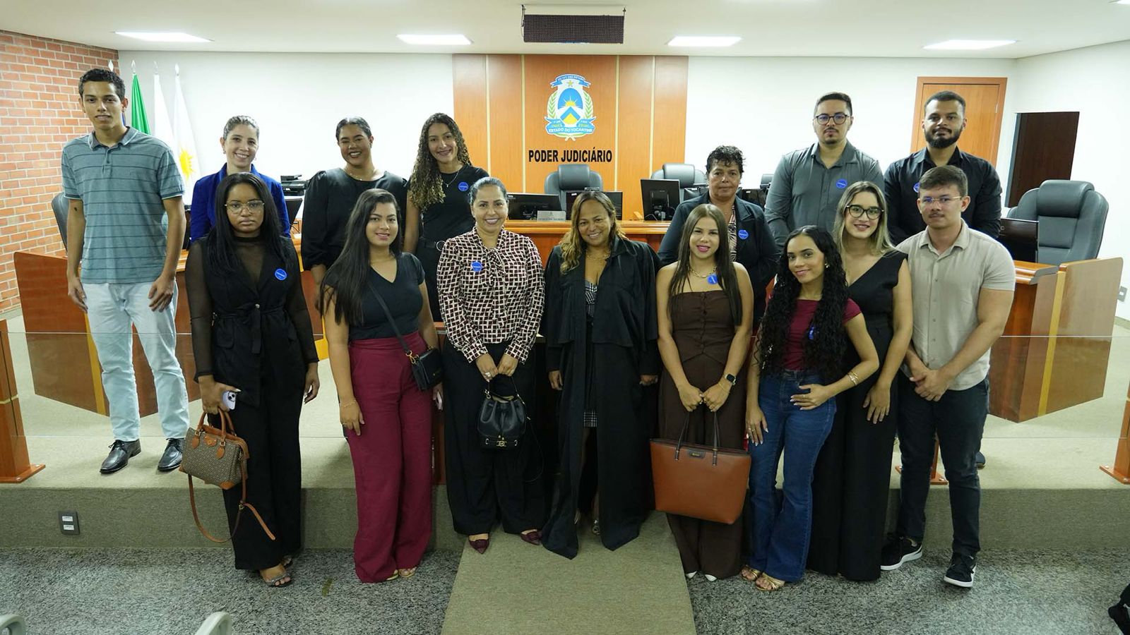 Homens e mulheres em pé posando pra foto dentro de sala de paredes brancas. Ao fundo, brasão do poder judiciário do Tocantins