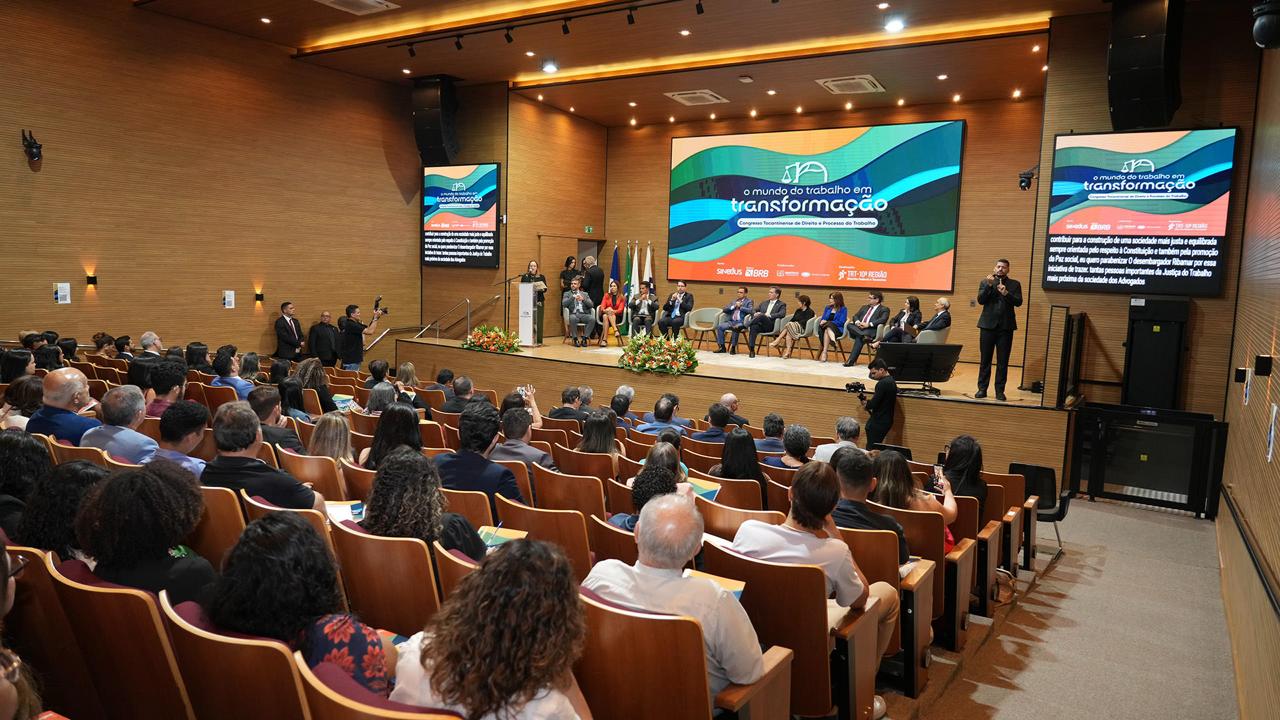 A imagem mostra o auditório amplo e moderno, com paredes de madeira clara e poltronas alaranjadas, está completamente ocupado por um público atento. No palco, cerca de dez pessoas estão sentadas em cadeiras, formando uma mesa de honra. À esquerda, uma mulher fala ao púlpito. Atrás dela, três bandeiras e um grande painel digital exibem o tema do evento: “O mundo do trabalho em transformação – Congresso Tocantinense de Direito e Processo do Trabalho.”